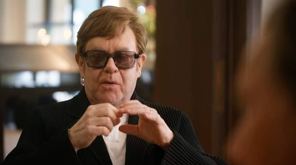 Dressed in a black pinstripe blazer and his signature embellished dark glasses, Elton John animatedly gestures with both hands as he recounts the story of his kneecap surgery to jeweler Theo Fennell in a scene from the documentary short "Elton John — Touched By Gold". | Source: YouTube/World Gold Council