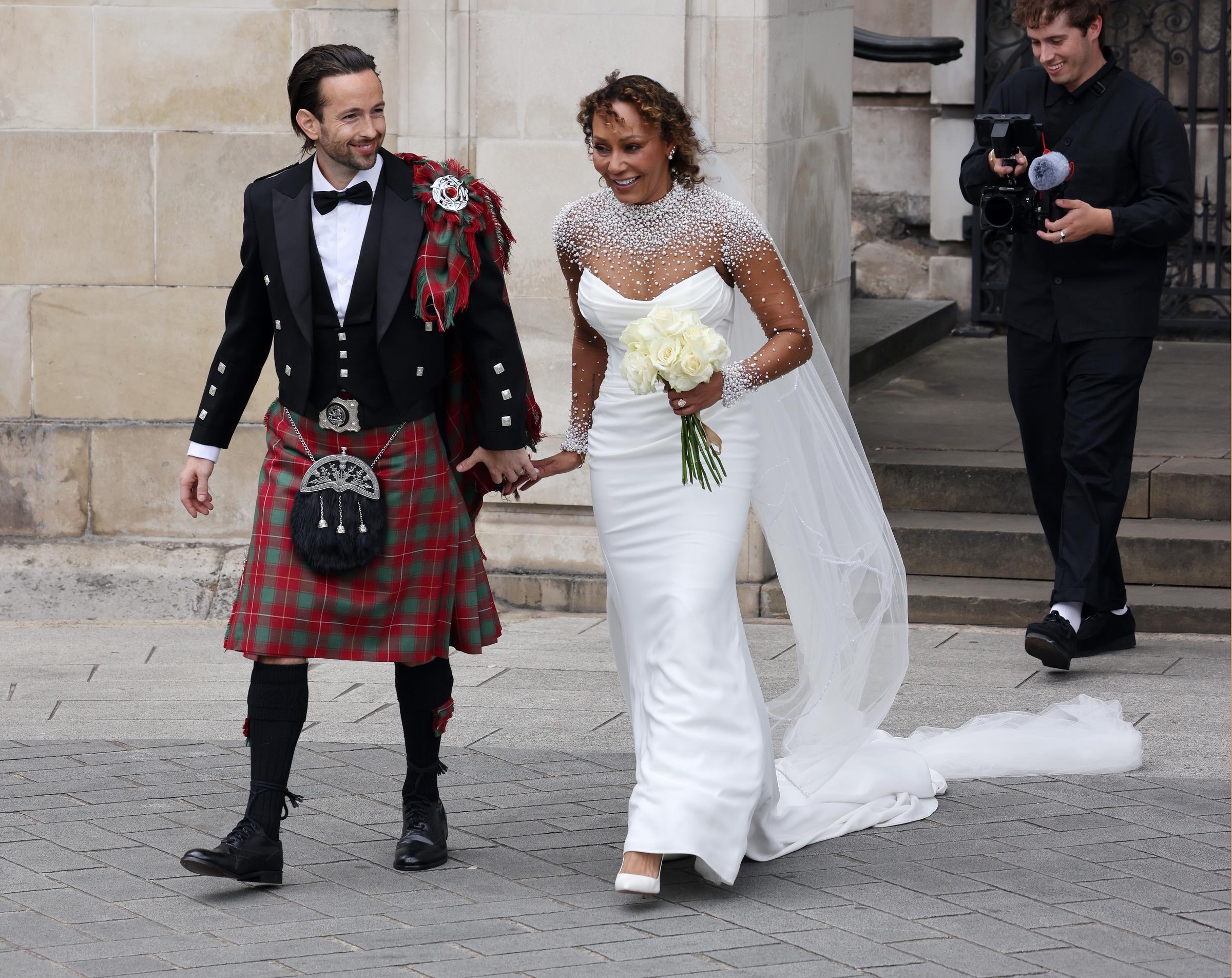 Captured in a full-length frame, Mel B and Rory McPhee make their exit from St. Paul’s Cathedral. Hand in hand, they smile broadly, their wedding attire gleaming under the daylight. The grandeur of the cathedral behind them contrasts beautifully with the intimacy of their shared moment.