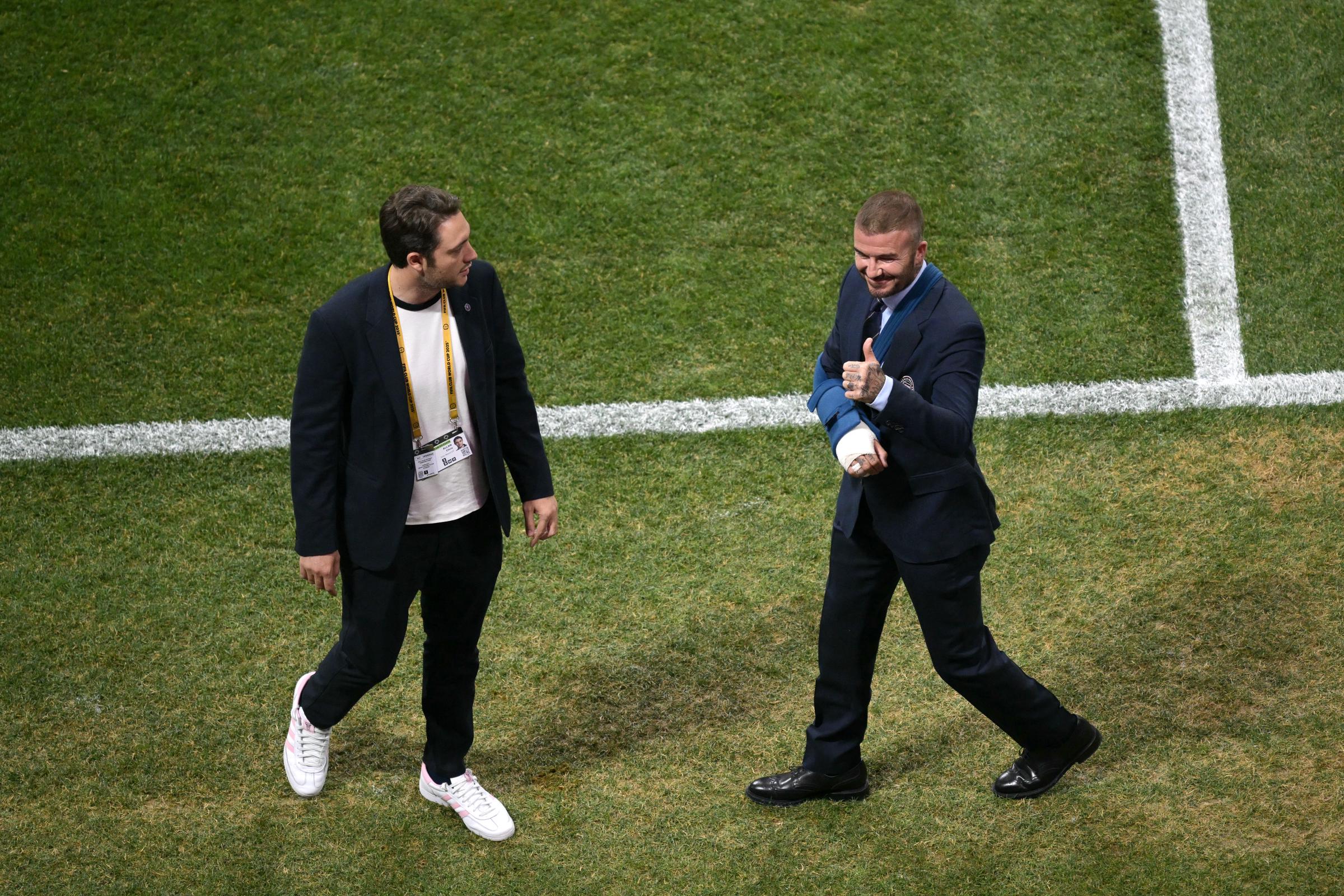 David Beckham walks across the field at Mercedes-Benz Stadium, flashing a thumbs-up as he approaches a colleague. His right arm is wrapped in a sling, but he appears upbeat and at ease.