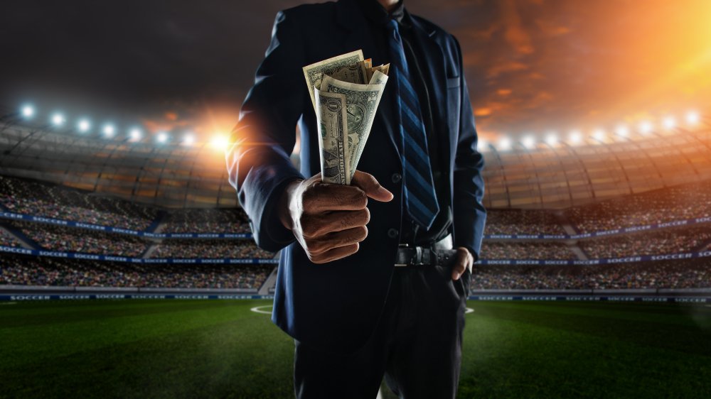 empresario sostiene una gran cantidad de billetes en el estadio de fútbol en segundo plano. I Foto: Shutterstock empresario sostiene una gran cantidad de billetes en el estadio de fútbol en segundo plano. I Foto: Shutterstock