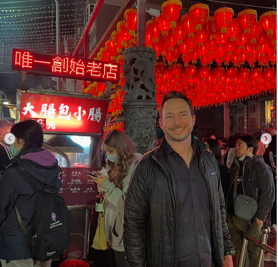 Matthew Mitchell smiles beneath glowing red lanterns while exploring a bustling night market lined with bright neon signs and crowded food stalls. | Source: Instagram/thy_travelers