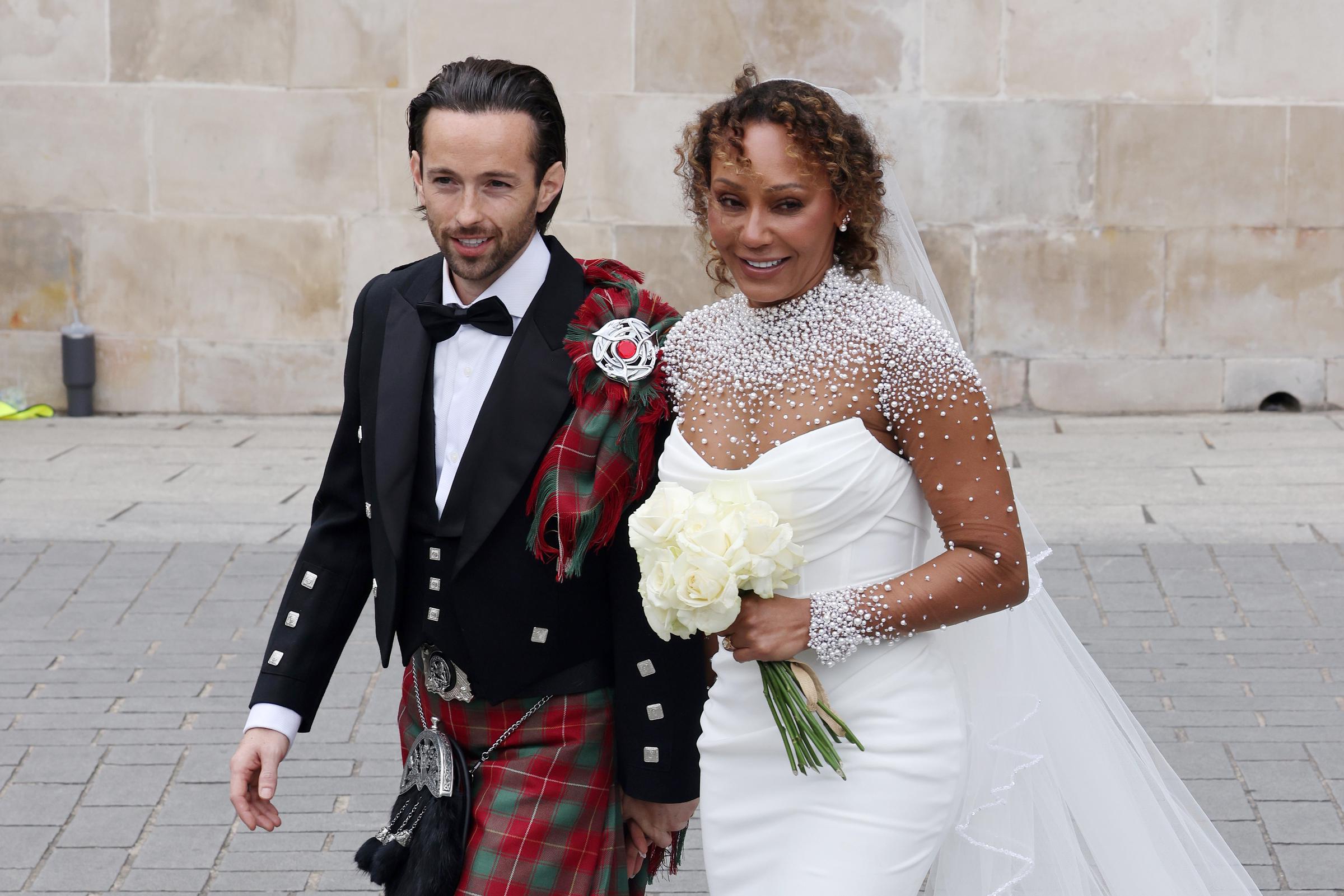 In a medium shot, newlyweds Mel B and Rory McPhee smile warmly as they depart St. Paul’s Cathedral. Surrounded by well-wishers, the couple radiates happiness, marking the joyful close of their wedding ceremony and the beginning of their life together.