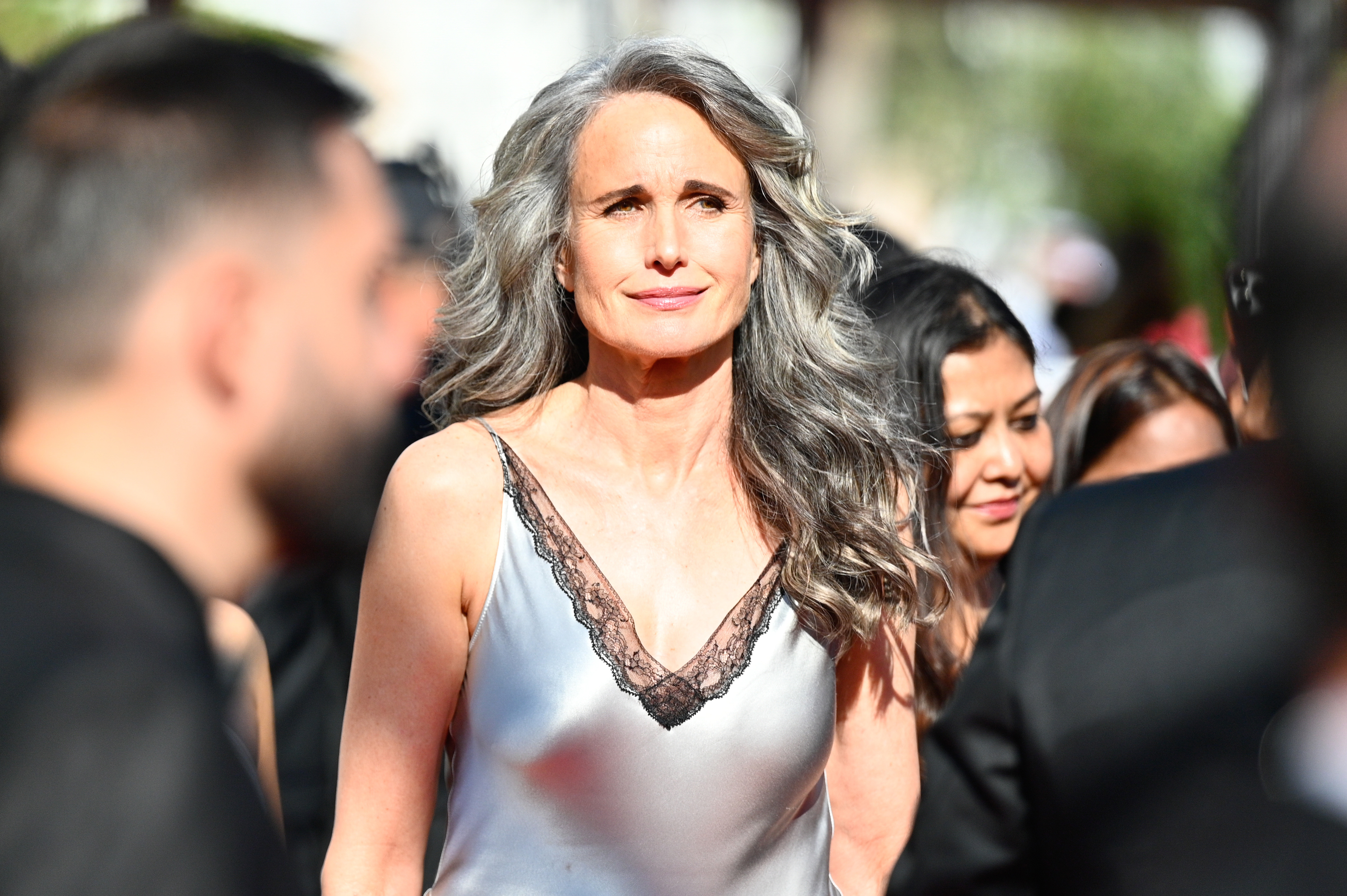 Among the crowd, Andie MacDowell stands out as sunlight illuminates her silver gown at the Cannes Film Festival. She looks ahead with a calm expression while guests gather behind her.