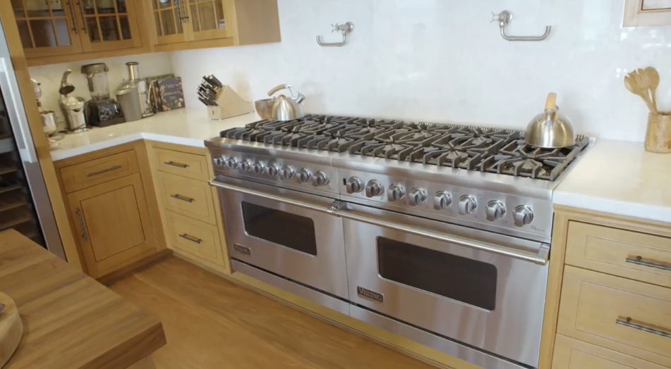 A expansive professional-grade Viking double-oven range serves as the focal point of the main kitchen, flanked by clean white countertops and polished light wood drawers. Polished kettles sit atop the massive gas cooktop, with wall-mounted pot fillers providing convenient access right above the cooking surface. | Source: YouTube/Architectural Digest