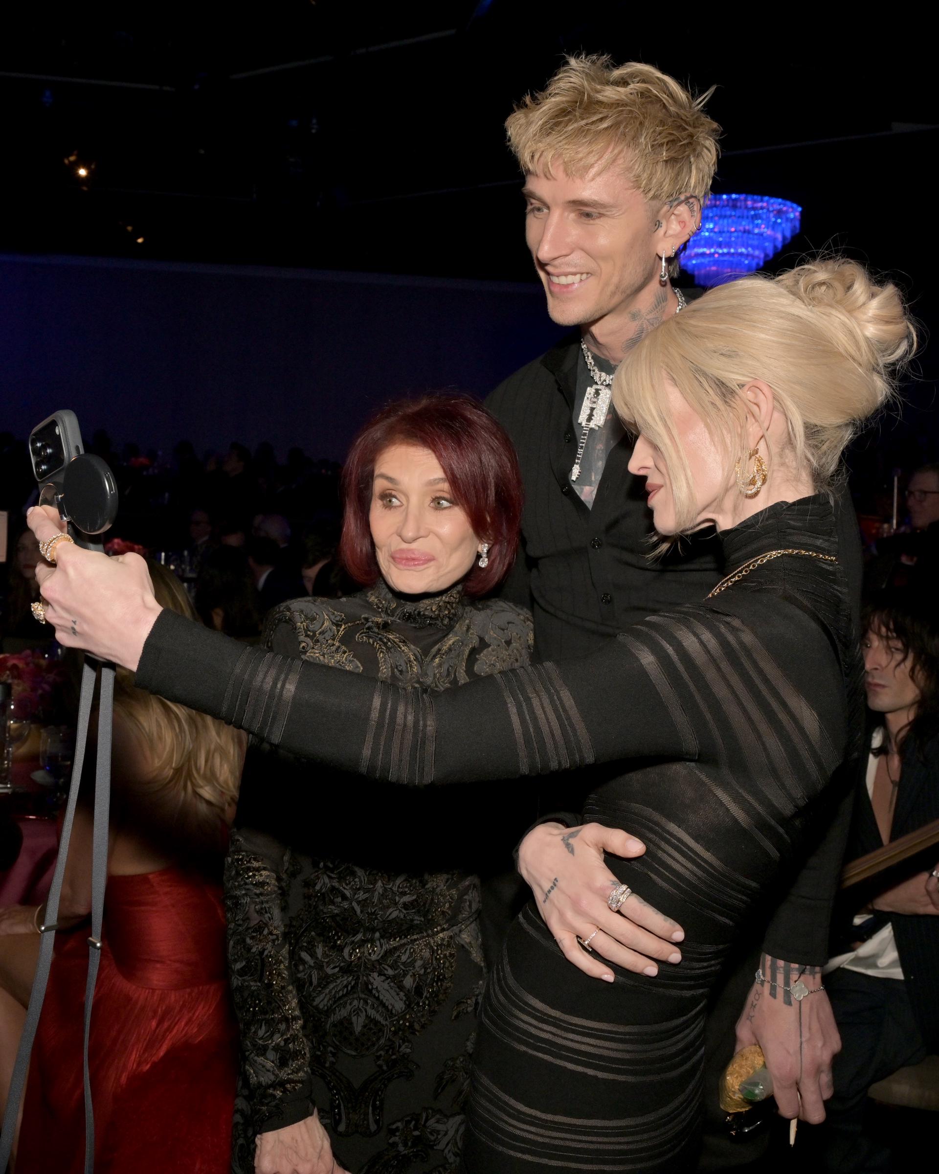 Sharon and Kelly Osbourne lean in alongside Machine Gun Kelly as they pause for a selfie inside the Beverly Hilton during the 68th Grammy Awards pre-Grammy gala on January 31, 2026, in Los Angeles. Surrounded by fellow industry figures, the trio appear relaxed and engaged in the moment, offering a candid glimpse into the evening.