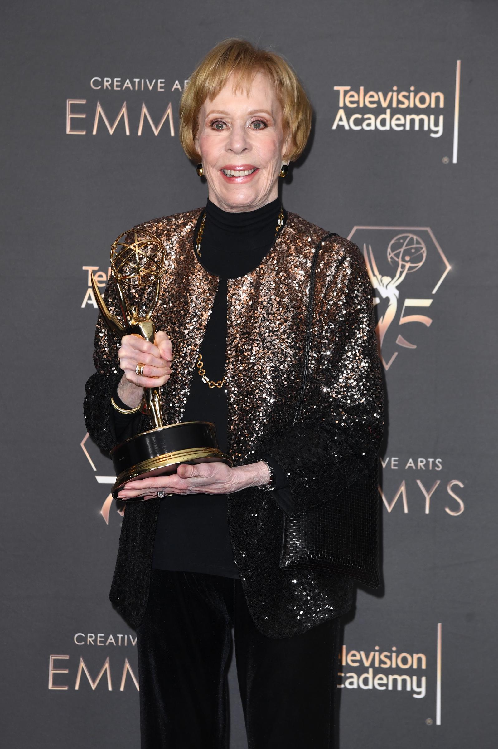 She poses with an award in hand, dressed in a black turtleneck top and a bronze sequined jacket, paired with dark trousers. A gold chain necklace and matching earrings complete the look. She stands against a step-and-repeat backdrop, holding the trophy at chest level while facing forward.