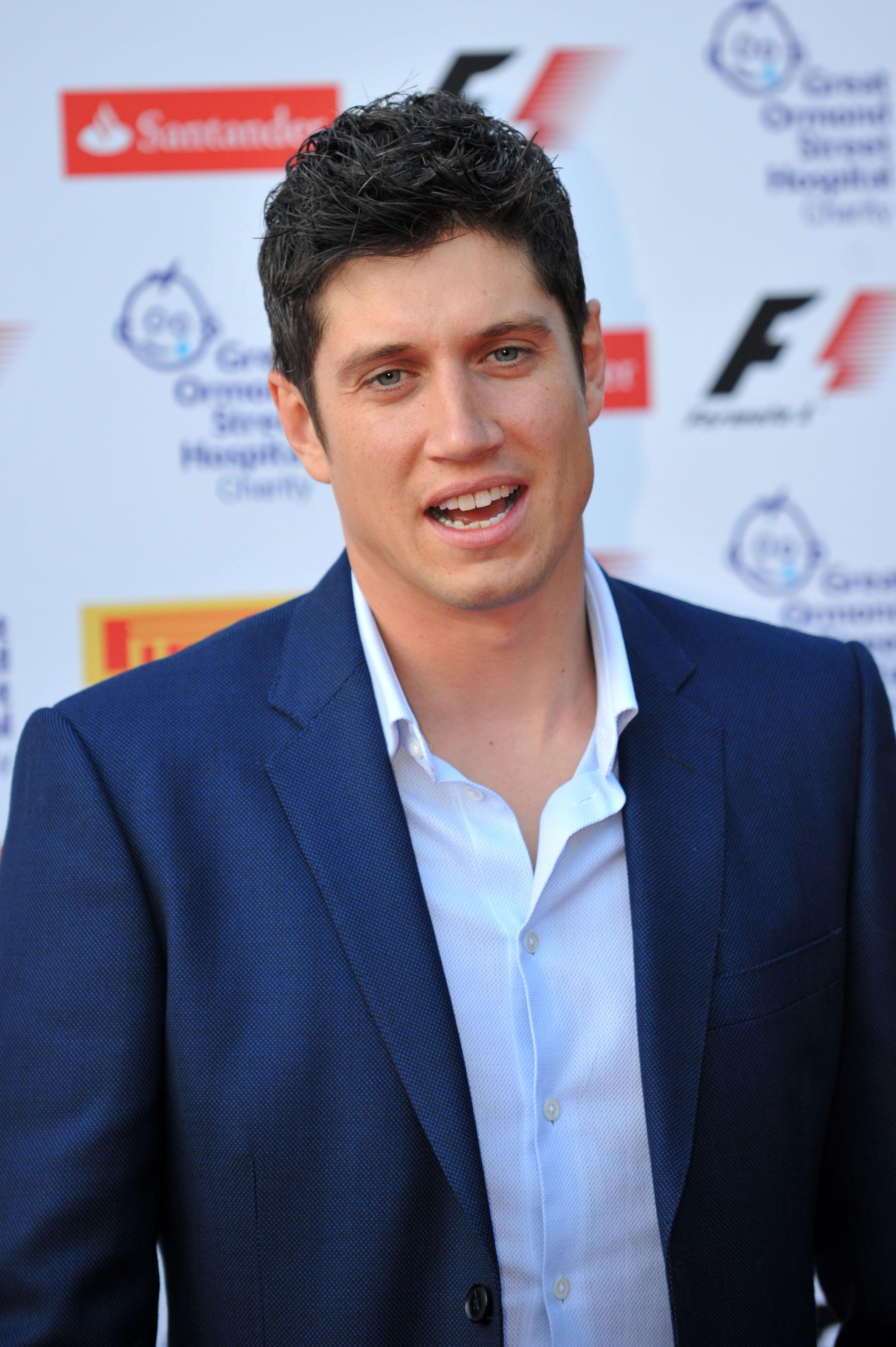 Vernon Kay sports a navy blazer over an open-collar white shirt, mid-sentence with a lively expression as he faces the camera.