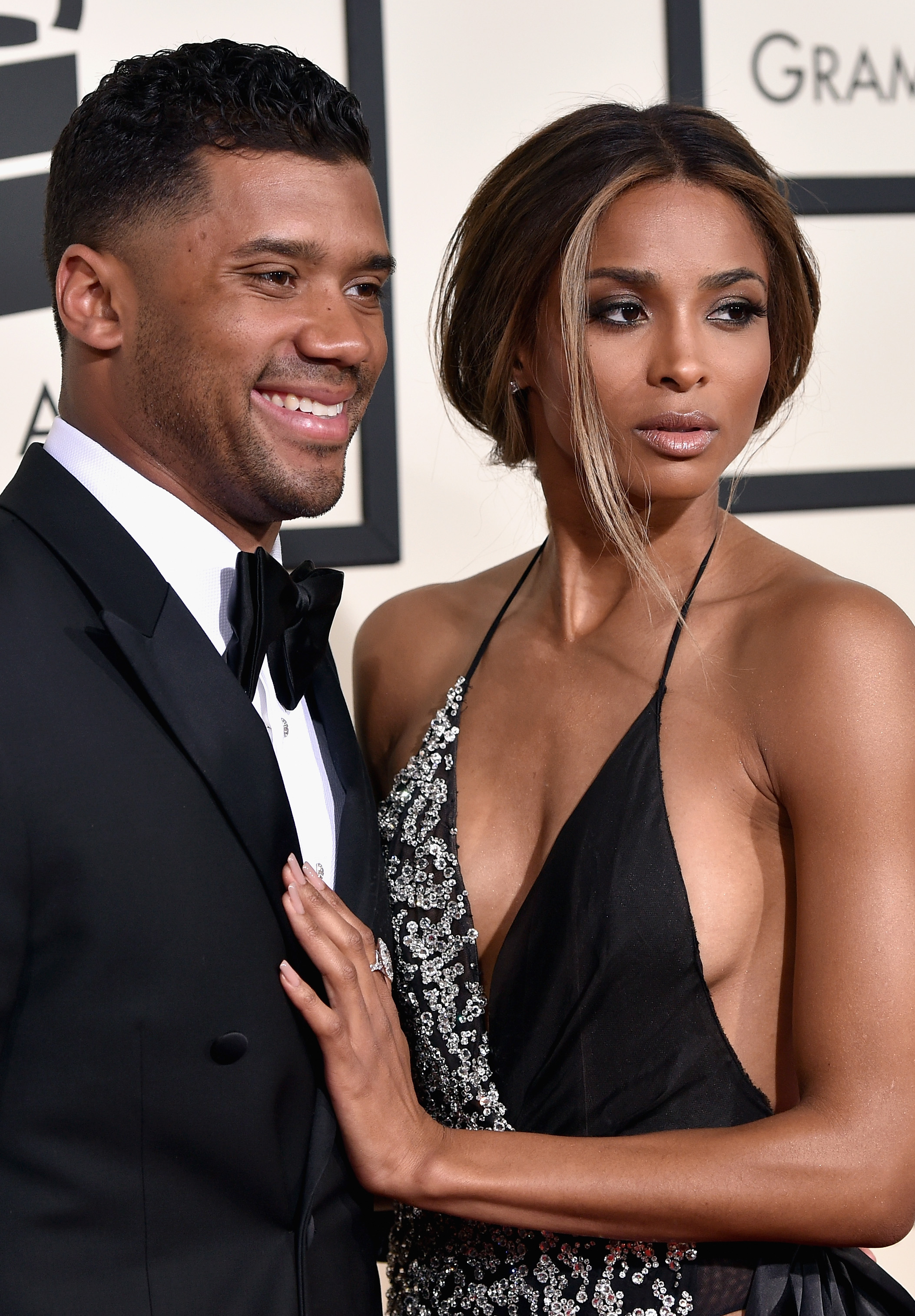 Russell Wilson, dressed in a classic tuxedo, smiles beside Ciara, who stuns in a sleek black gown with a refined, elegant finish. She turns slightly with a poised, striking expression, adding a touch of drama to the otherwise composed moment.