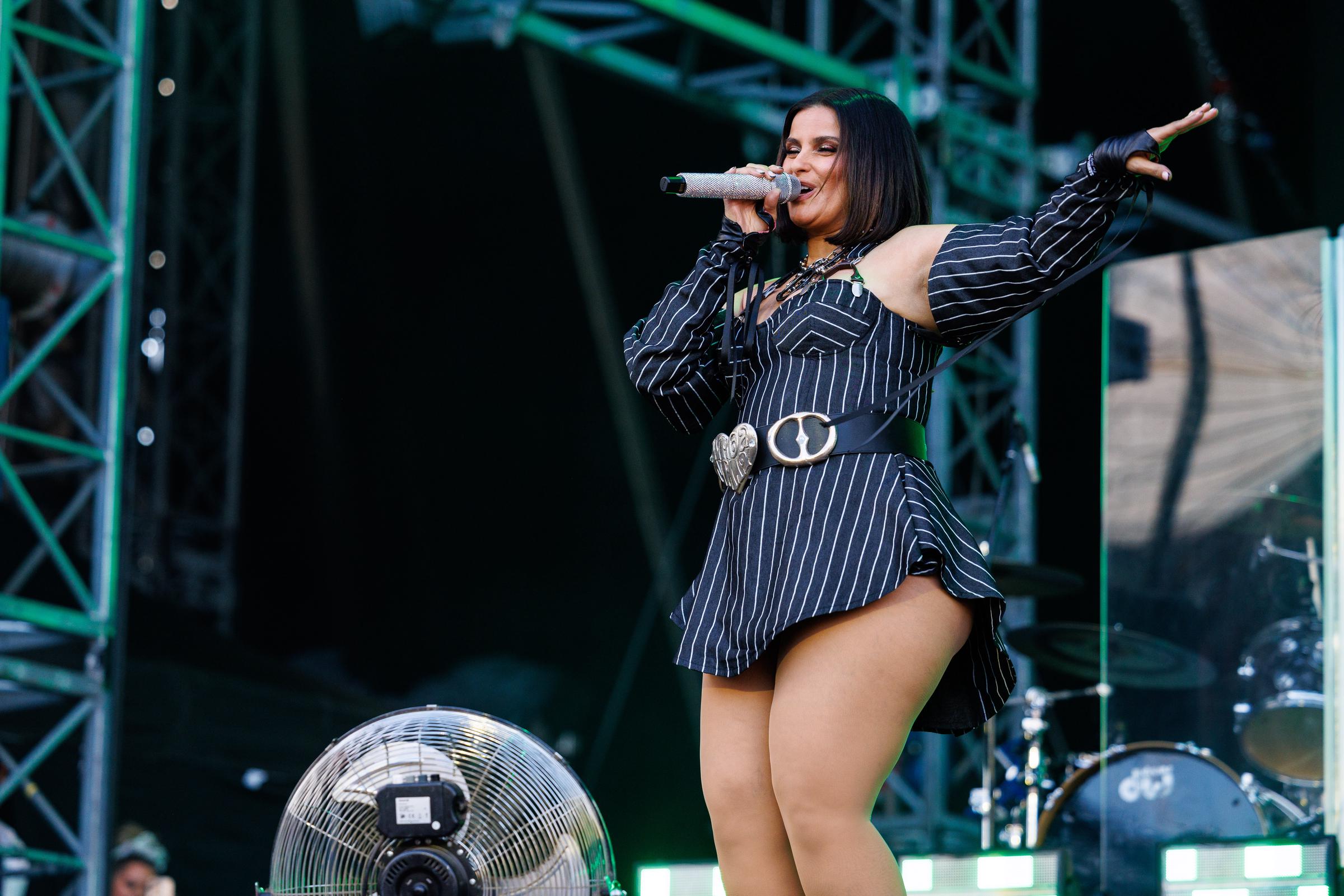 Furtado performs on stage in a black and white pinstriped one-shoulder mini dress with a dramatic puffed sleeve on one side. She accessorizes with a statement silver belt buckle at the waist, a chunky chain necklace, and her hair is styled sleek and straight in a shoulder-length bob.