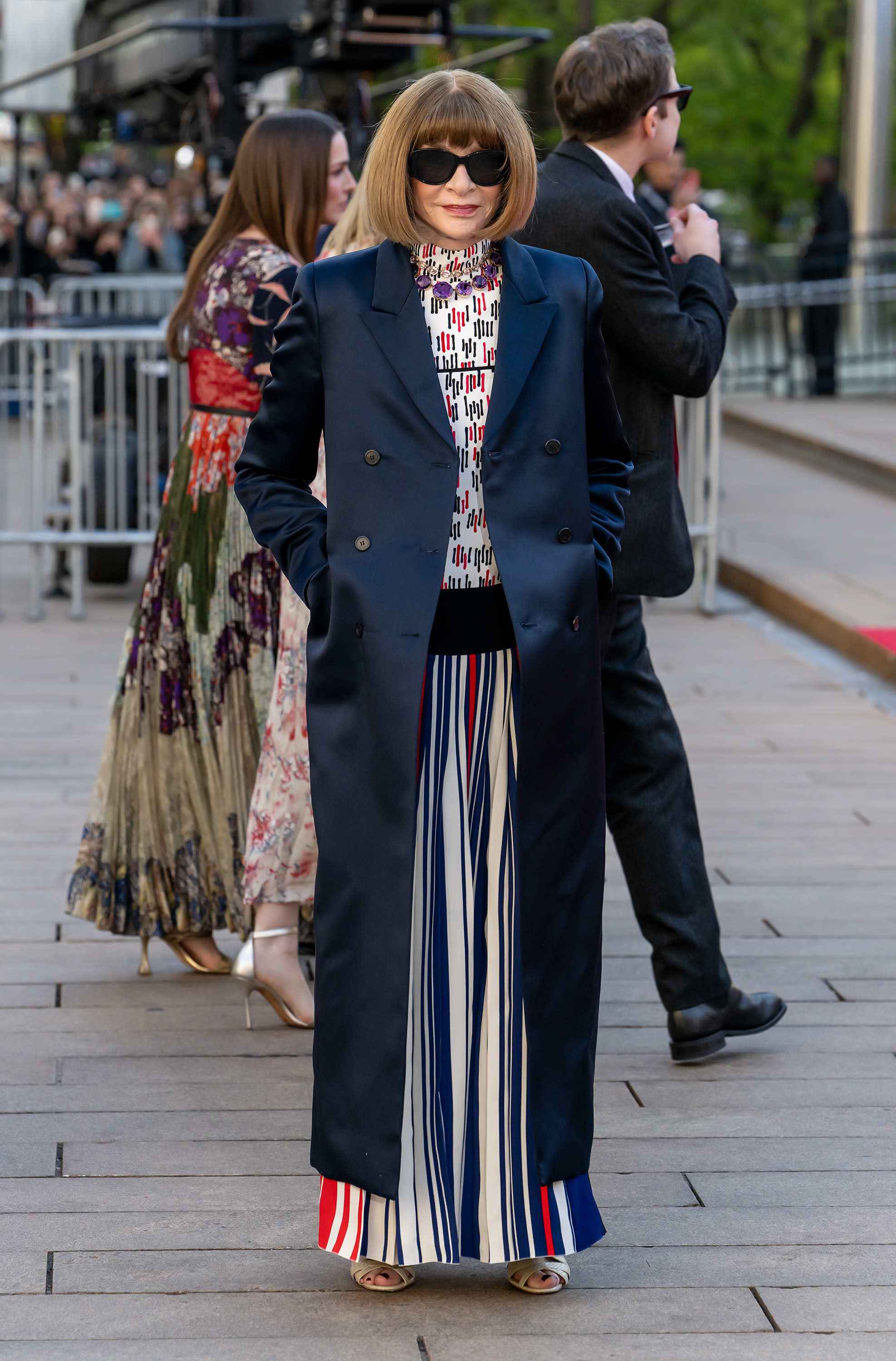 Anna Wintour arrives off the main carpet in a navy double-breasted longline blazer over a geometric print turtleneck top, wide-leg red, white, and navy striped palazzo trousers, a bold amethyst necklace, and gold open-toe heels.