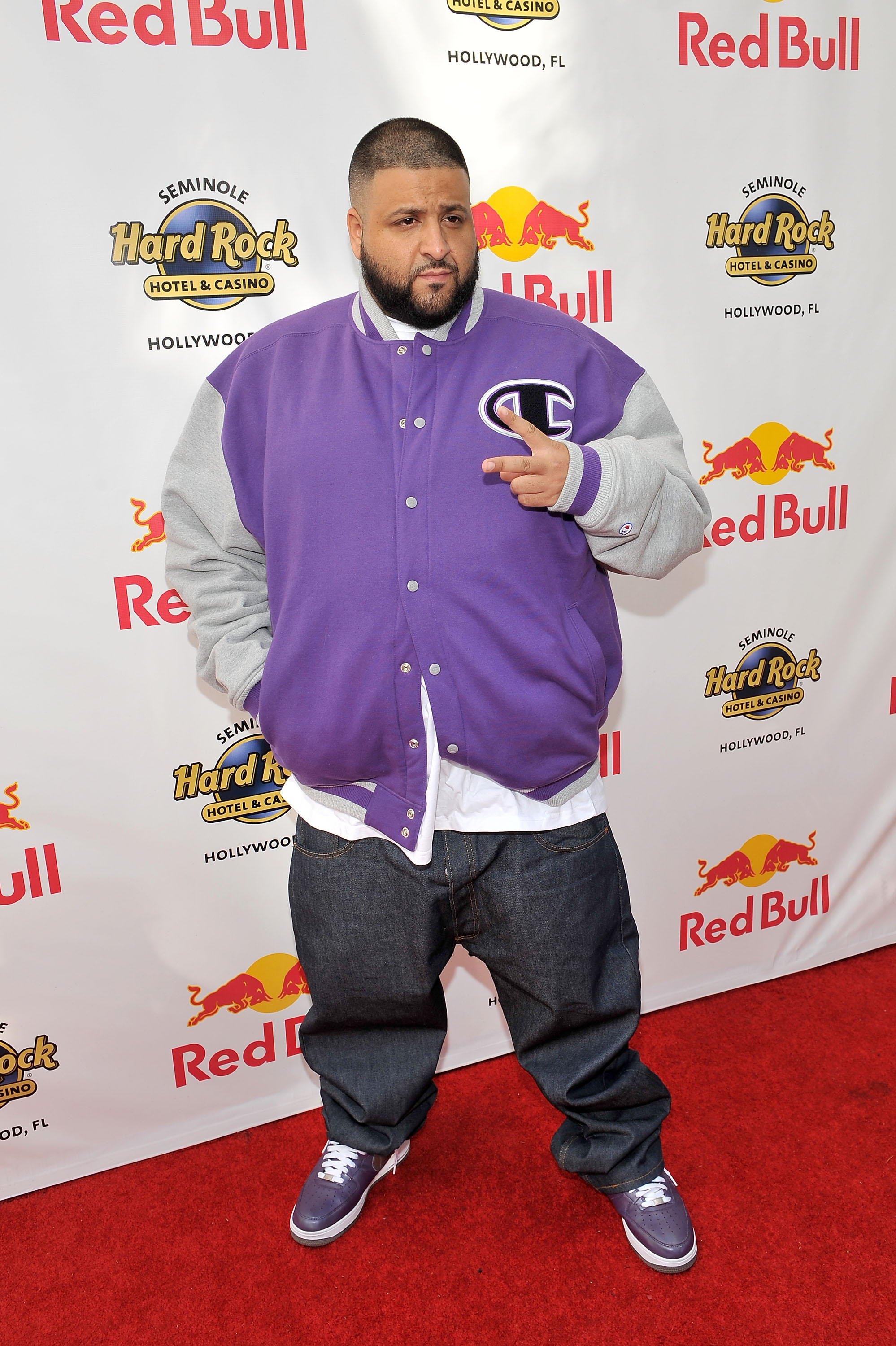 On the red carpet, DJ Khaled wears a purple varsity jacket over a white tee, paired with loose denim and coordinating sneakers while flashing a casual peace sign in front of a branded event wall.