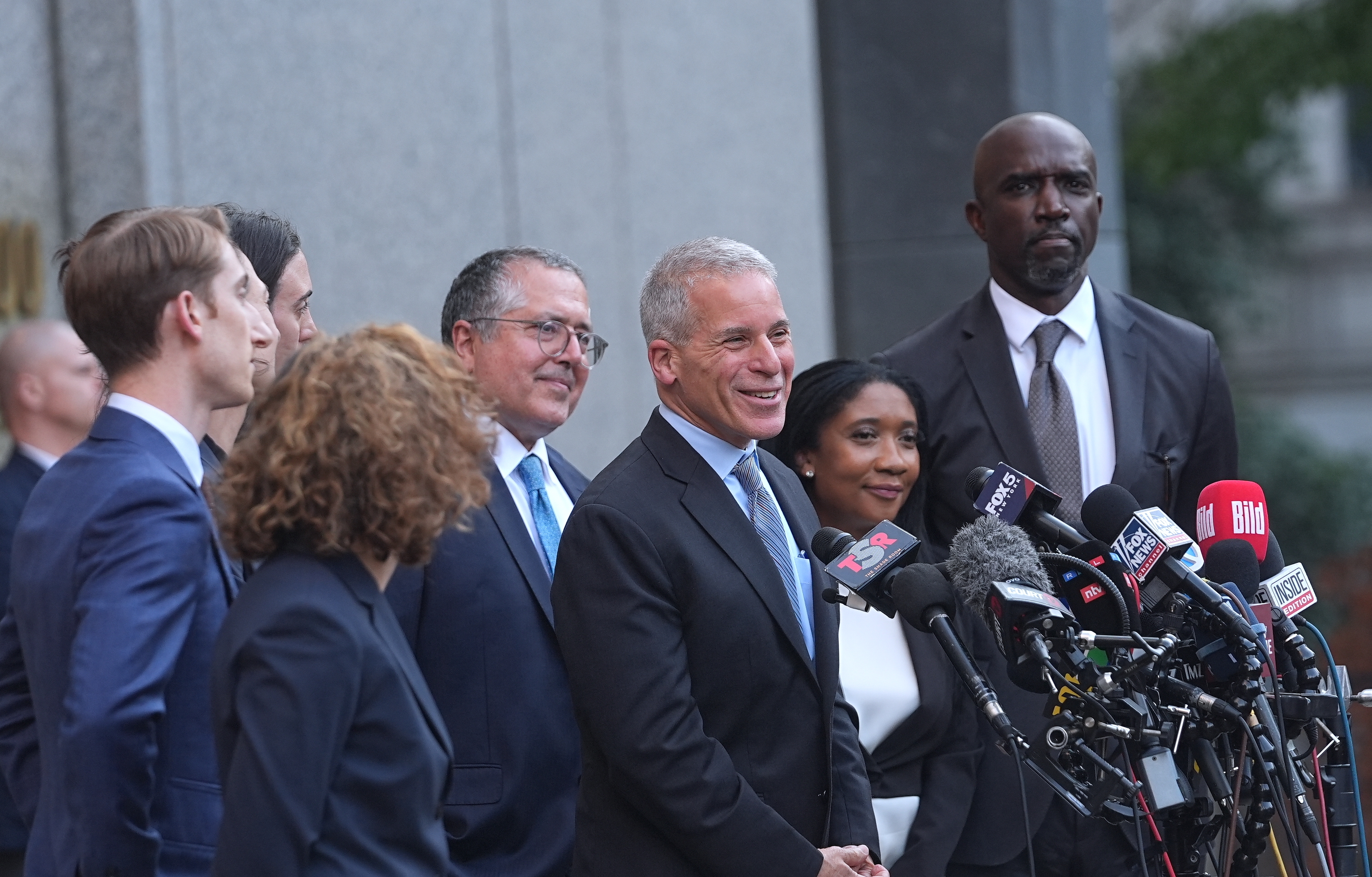Attorney Brian Steel leads the press conference with a wide smile, surrounded by fellow defense lawyers. Their expressions show relief as they address the media.
