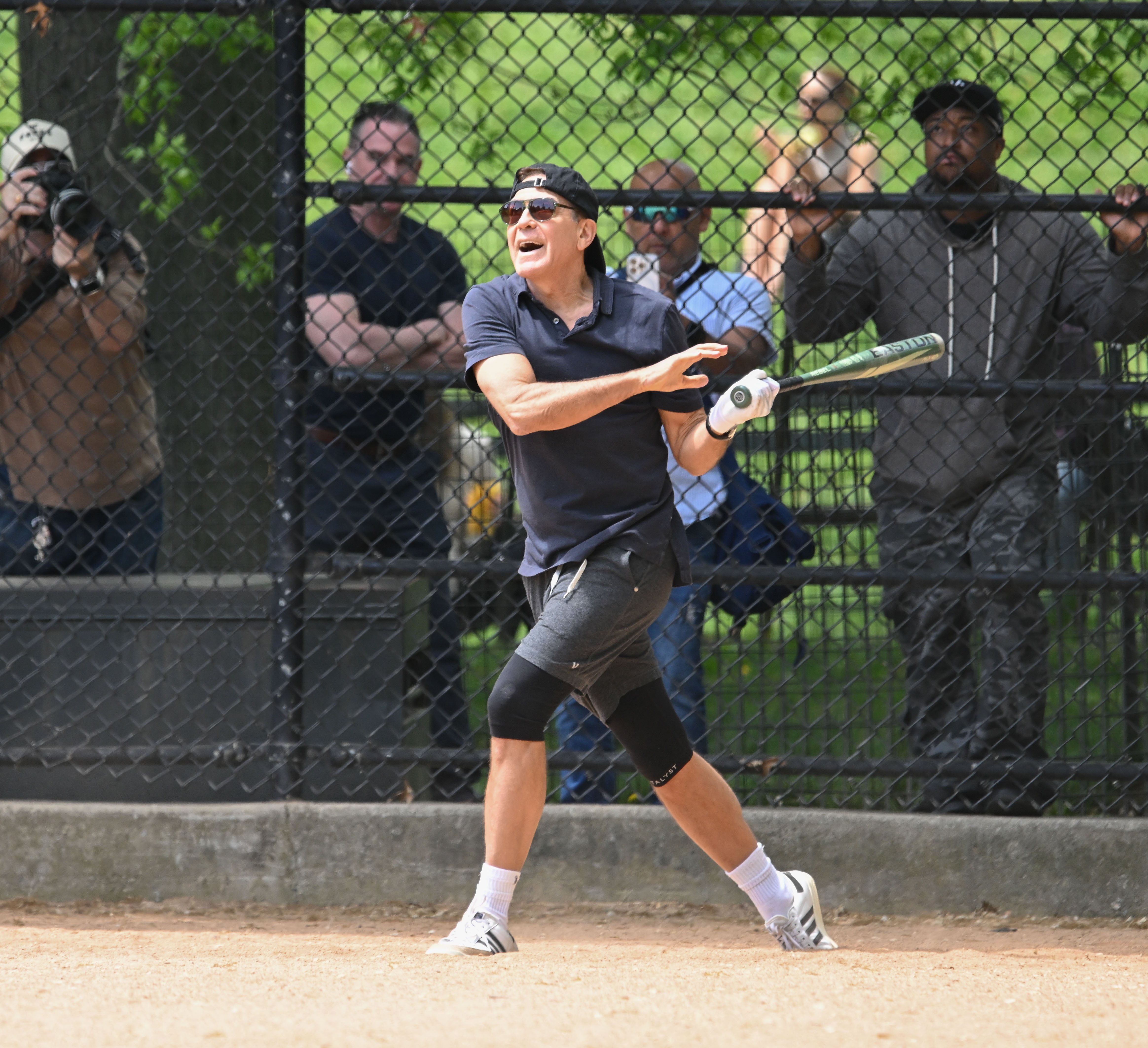 Clooney follows through on a swing, eyes tracking the ball with intensity and focus. His stance and expression capture the thrill of the moment, as spectators behind the fence look on, some cheering, others snapping photos.