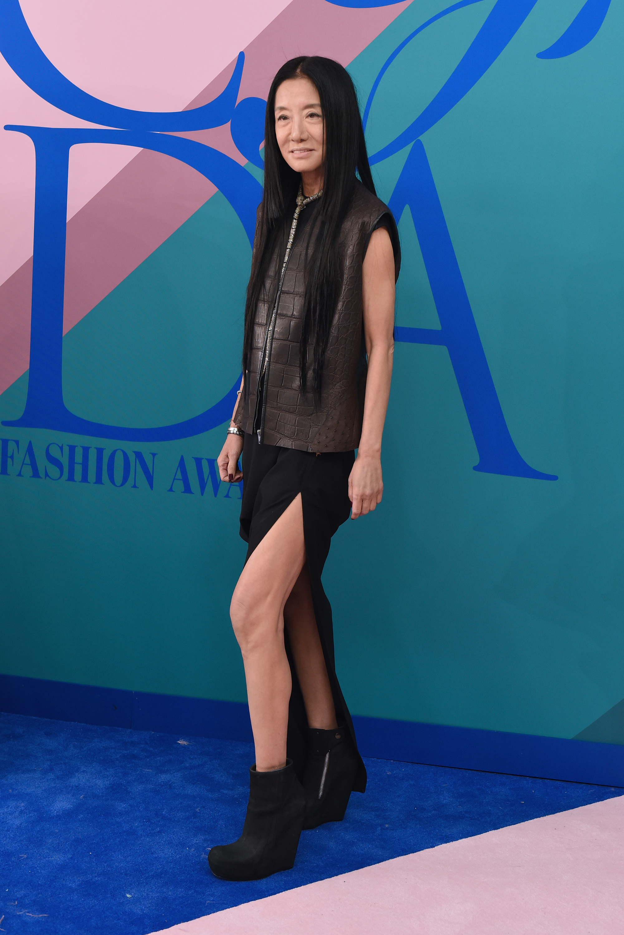 Vera Wang appears at the CFDA Fashion Awards in a black asymmetrical dress with a high slit, paired with a structured brown leather vest. She completes the edgy look with black booties and minimal accessories, letting the textures stand out.