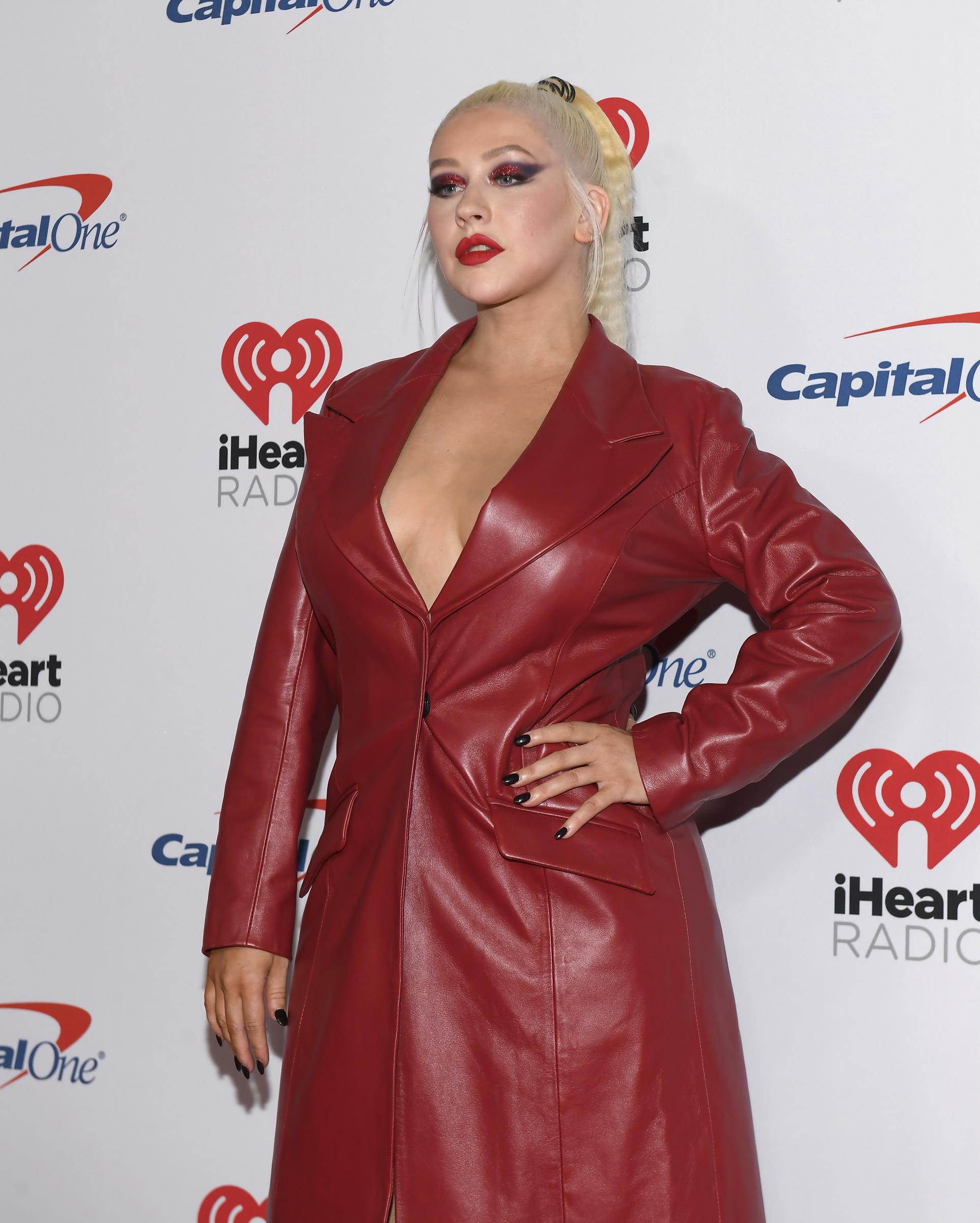 Christina Aguilera attends the 2019 iHeartRadio Music Festival at T-Mobile Arena in Las Vegas. She wears a bold red leather trench coat with a plunging neckline, paired with slicked-back platinum hair, crimson lipstick, and dramatic eye makeup, embodying her fierce and fearless style evolution.