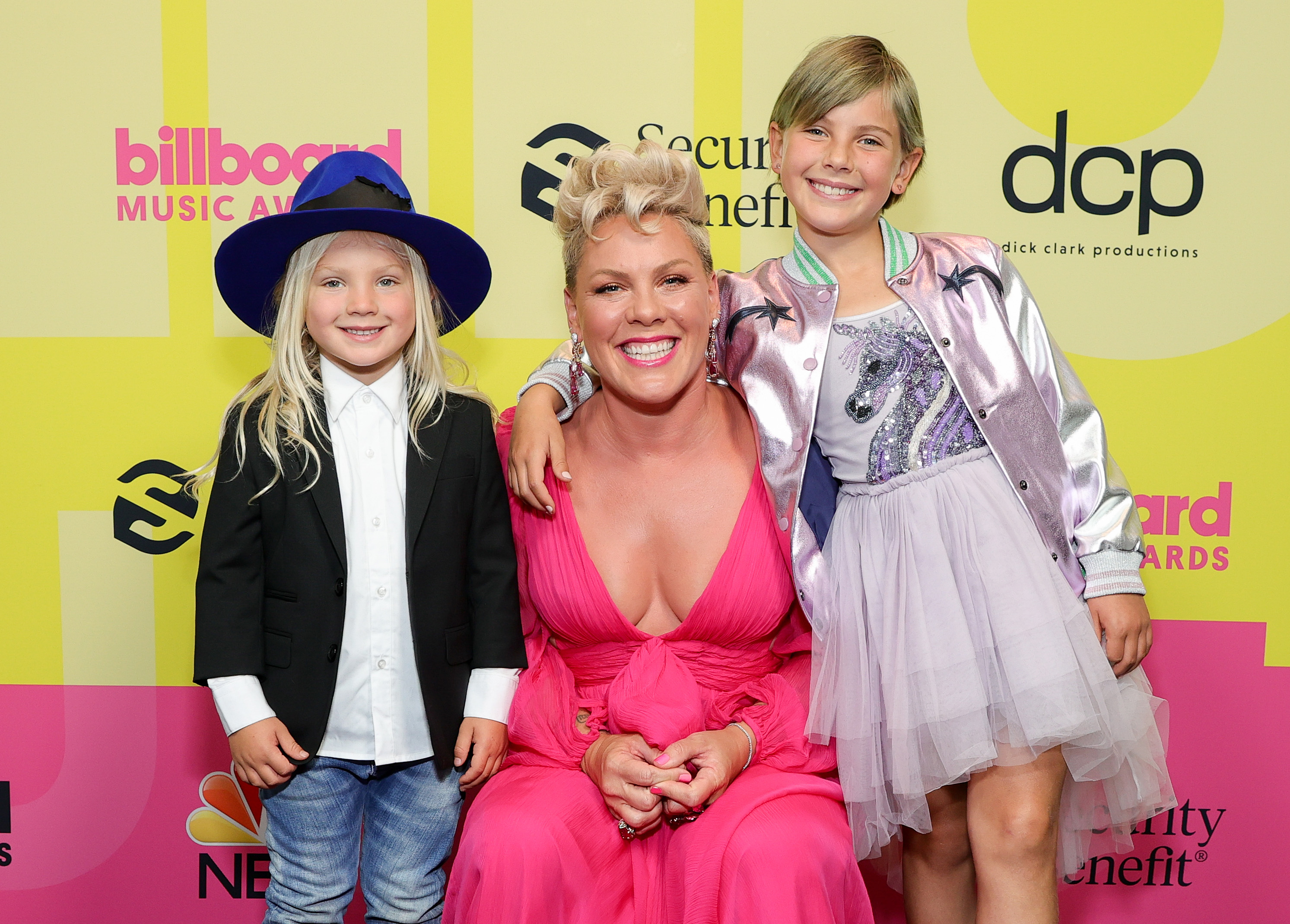 Jameson Moon Hart, Pink, and Willow Sage Hart pose backstage for the Billboard Music Awards. Pink smiles in a bright pink gown as Willow leans in wearing a lavender skirt and shiny jacket.
