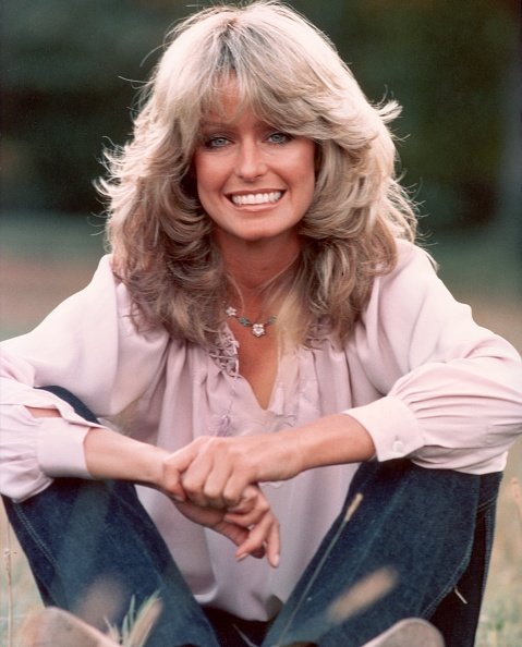 Undated photo of Farrah Fawcett smiling while sitting outdoors in blue jeans and a mauve blouse. | Photo: Getty Images
