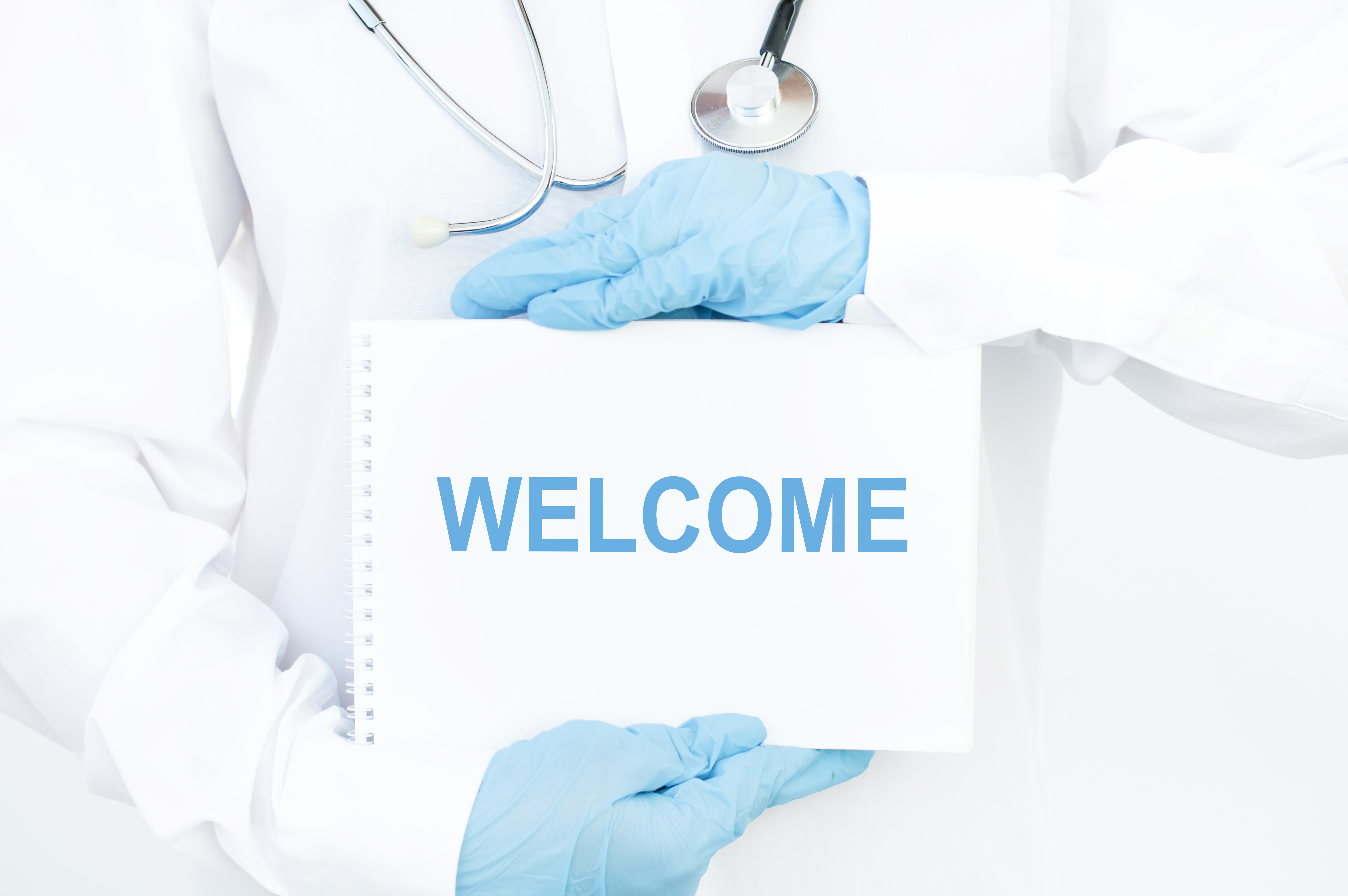 Arzt mit weißem Kittel und blauen Handschuhen hält ein "Willkommen"-Schild.|Quelle: Shutterstock