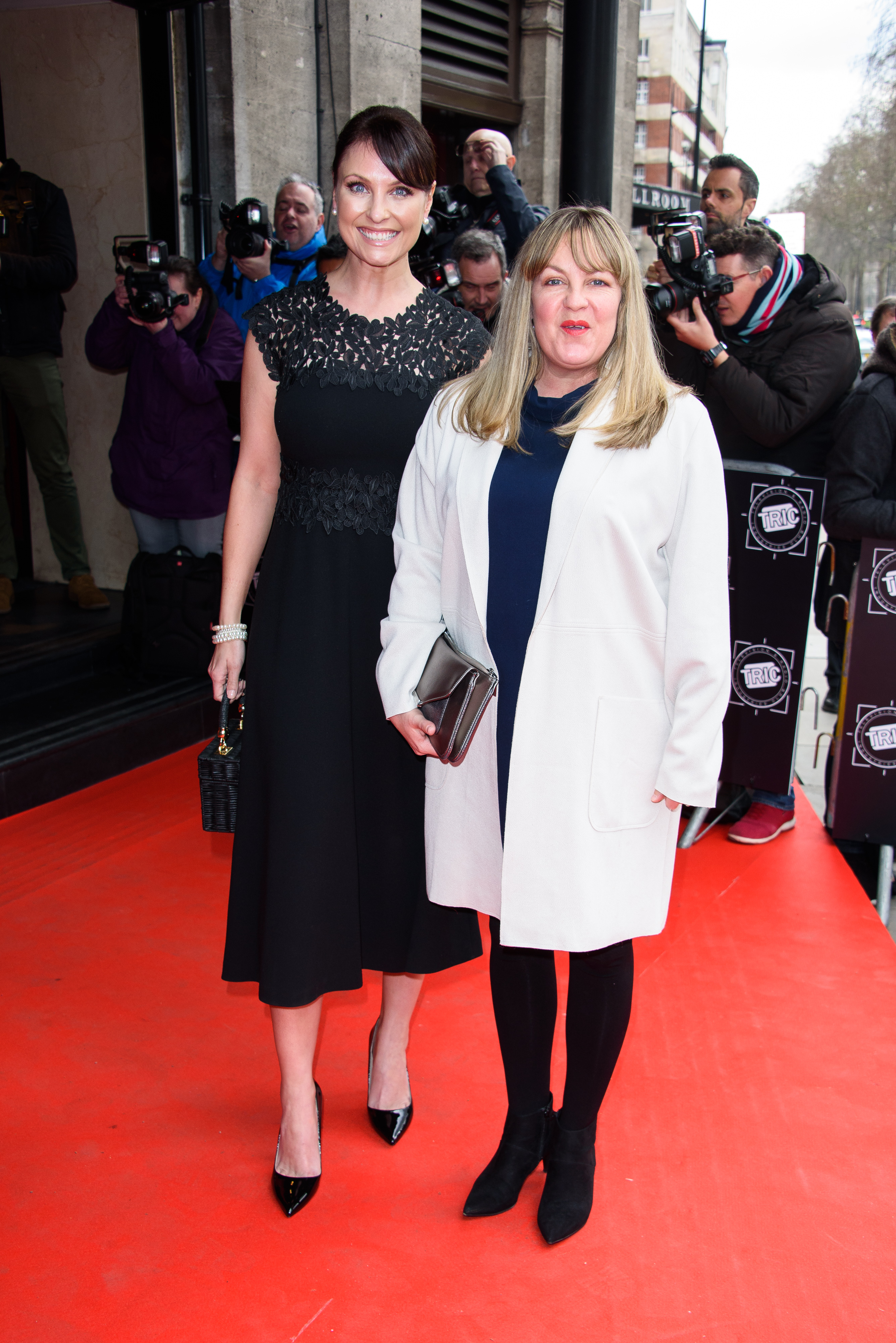 Emma Barton and Lorraine Stanley dazzle at the 2018 TRIC Awards, turning heads outside London's Grosvenor House Hotel on 13 March. The "EastEnders" duo strike a pose amid a flashbulb frenzy — Barton stuns in black lace while Stanley oozes confidence in a sharp white coat over navy.