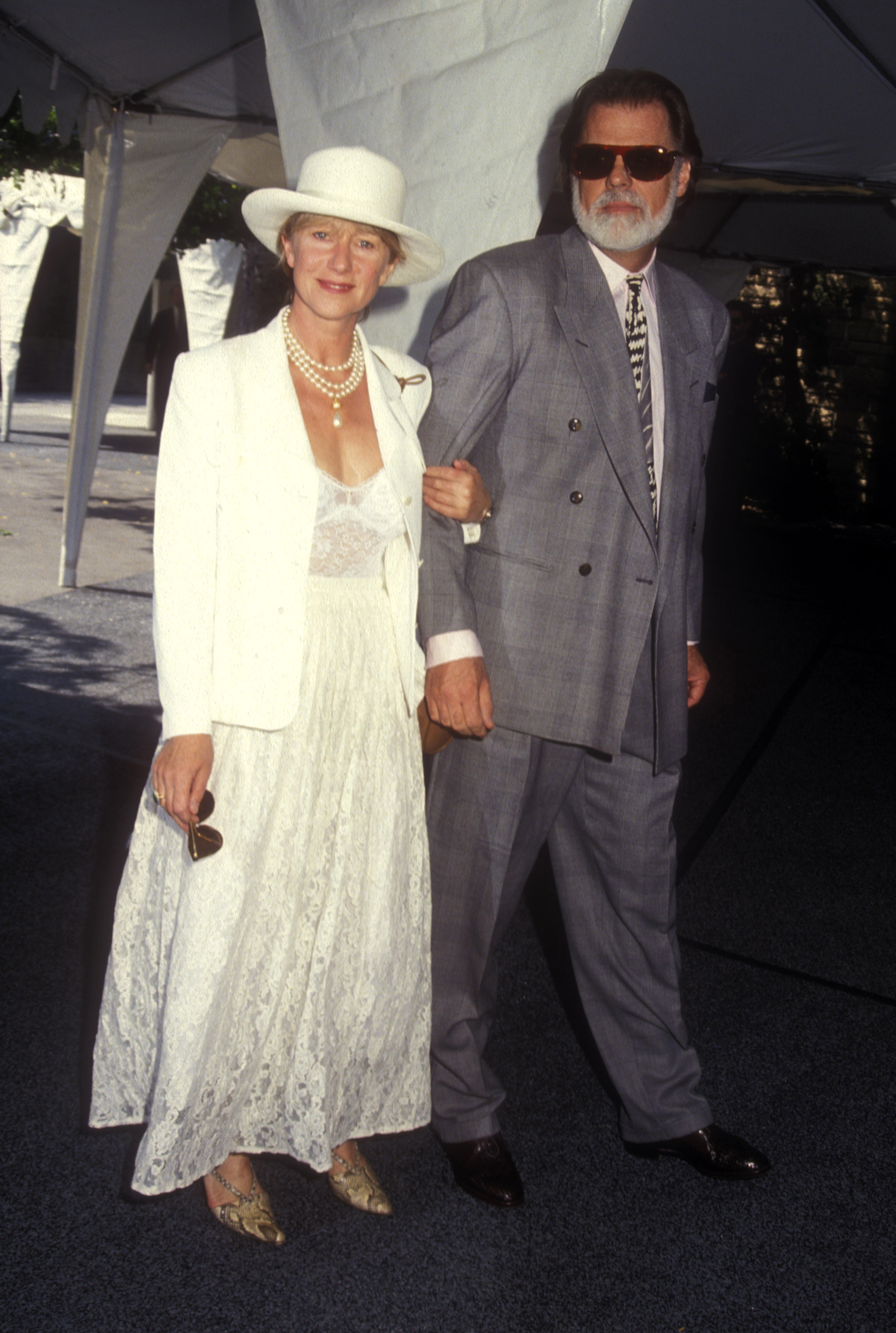 Helen Mirren arrives arm-in-arm with Taylor Hackford, dressed in a vintage-inspired white lace ensemble topped with a structured blazer and wide-brimmed hat, accessorized with layered pearls and gold heels, exuding classic elegance at a daytime event.
