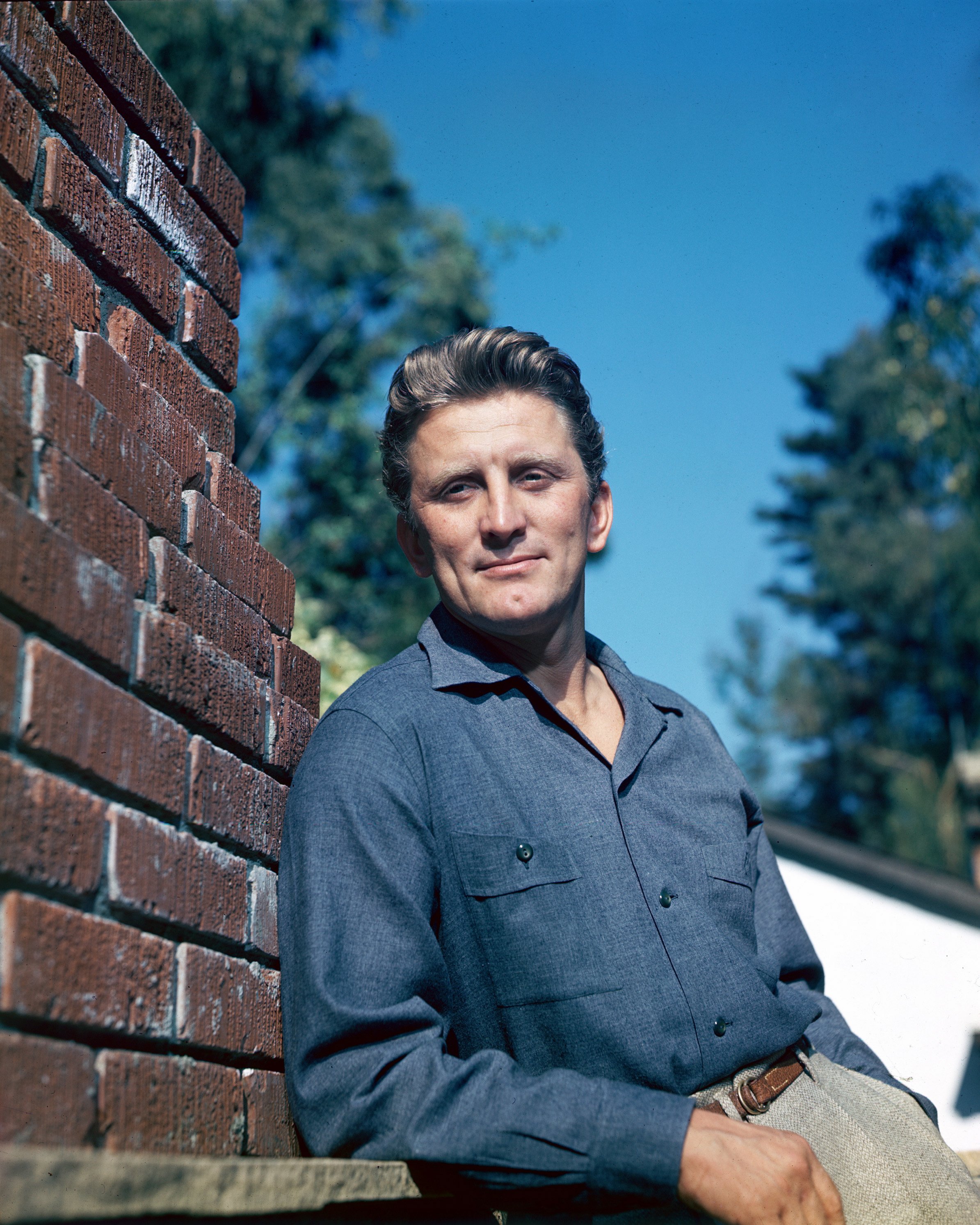 Kirk Douglas photographed leaning against the wall wearing a blue shirt paired with gray trousers in 1955. / Source: Getty Images