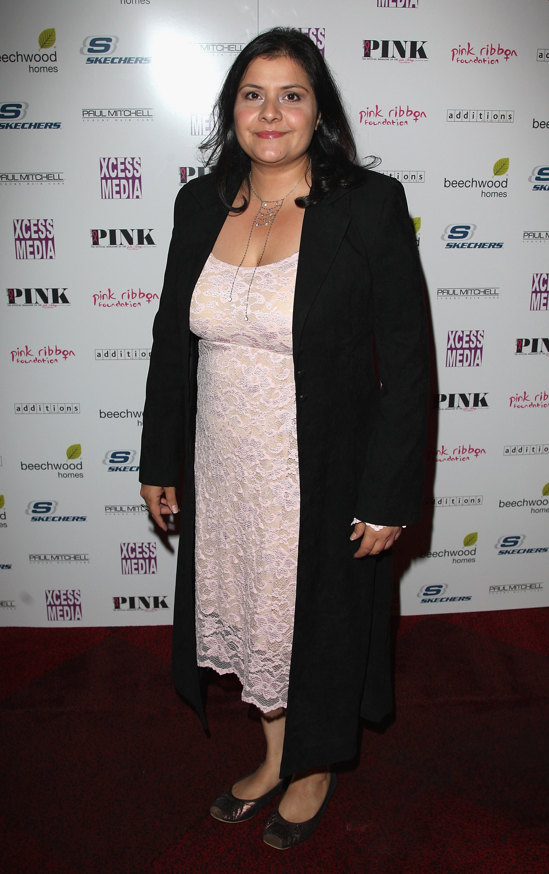 Nina Wadia stands full-length on the red carpet at the In The Pink Magazine Launch Party in London, October 2007, her dark hair loose around her shoulders. She wears a pale blush lace midi dress with a delicate floral pattern, layered beneath a long, open black coat, and completes the look with dark open-toed flats and a layered silver necklace.