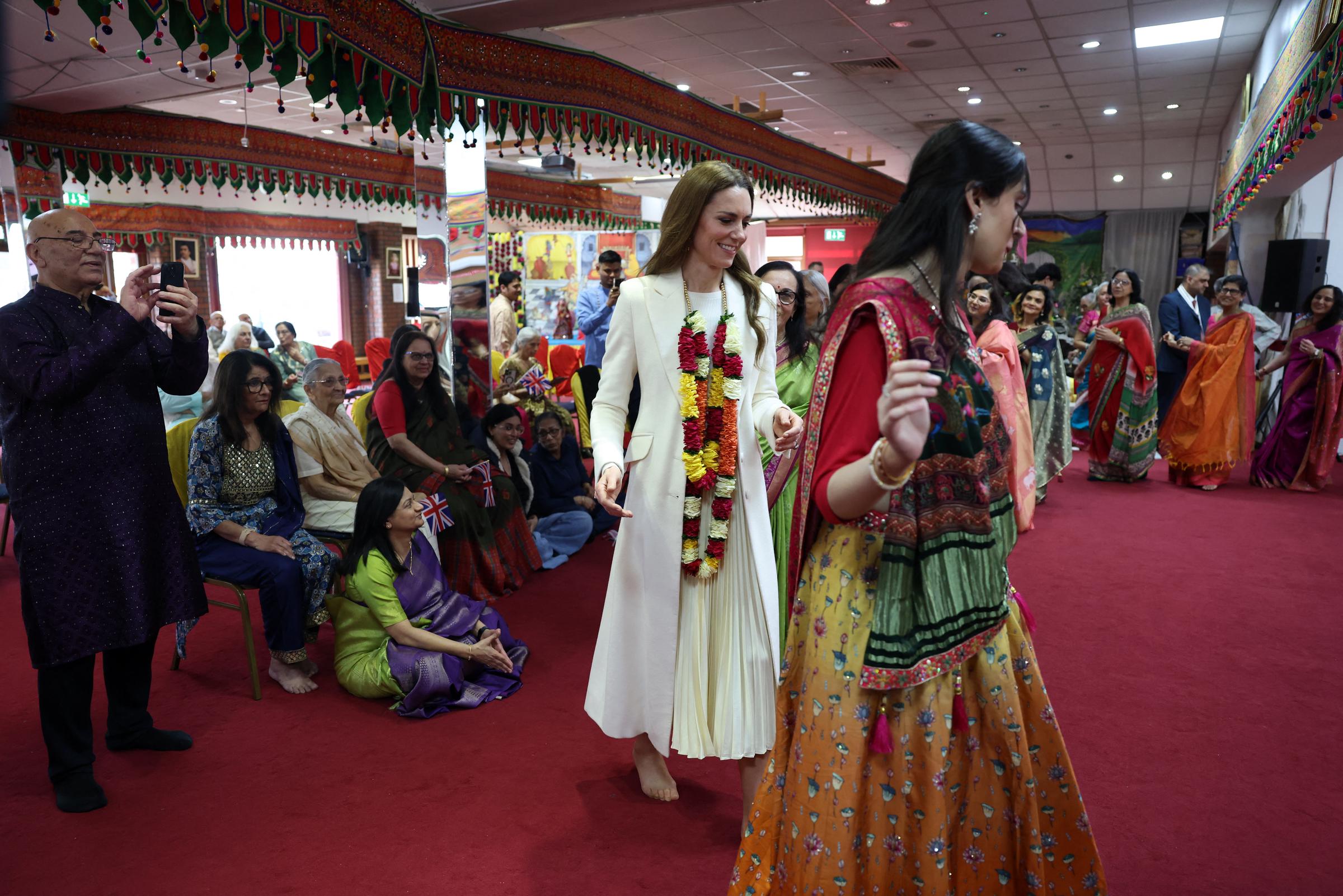 Catherine, Princess of Wales, joins participants during celebrations at the Shreeji Dham Haveli Hindu Temple in Leicester on March 5, 2026. Barefoot and wearing a floral garland presented during the visit, the Princess of Wales follows along with dancers moving through the hall, as spectators seated nearby watch and capture the moment on their phones.