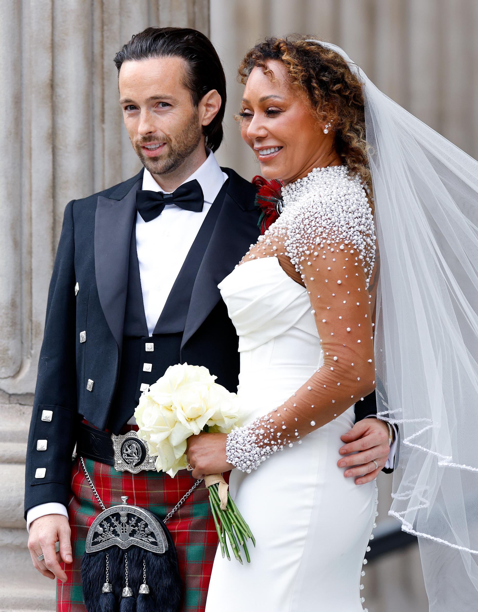 Mel B stands arm in arm with her new husband, Rory McPhee. Dressed in a stunning white gown, she holds a bouquet of fresh blooms, her expression radiant. Beside her, McPhee stands proudly, the pair capturing a moment of joy and calm amid the excitement of their wedding day.