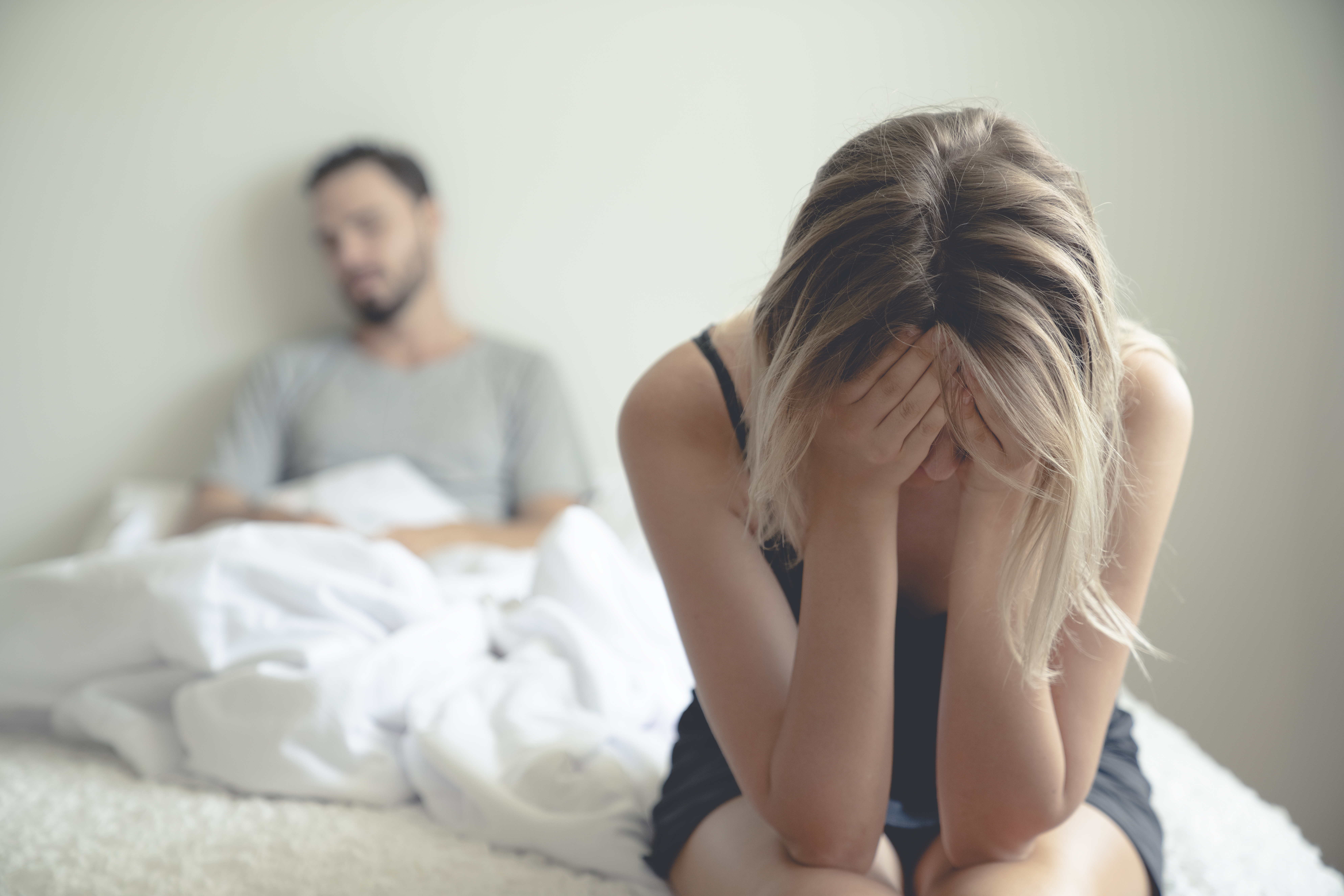 Eine frustrierte und traurige Frau, die auf einem Bett sitzt und mit den Händen ihr Gesicht bedeckt, während im Hintergrund ein Mann unter der Decke zu sehen ist | Quelle: Getty Images