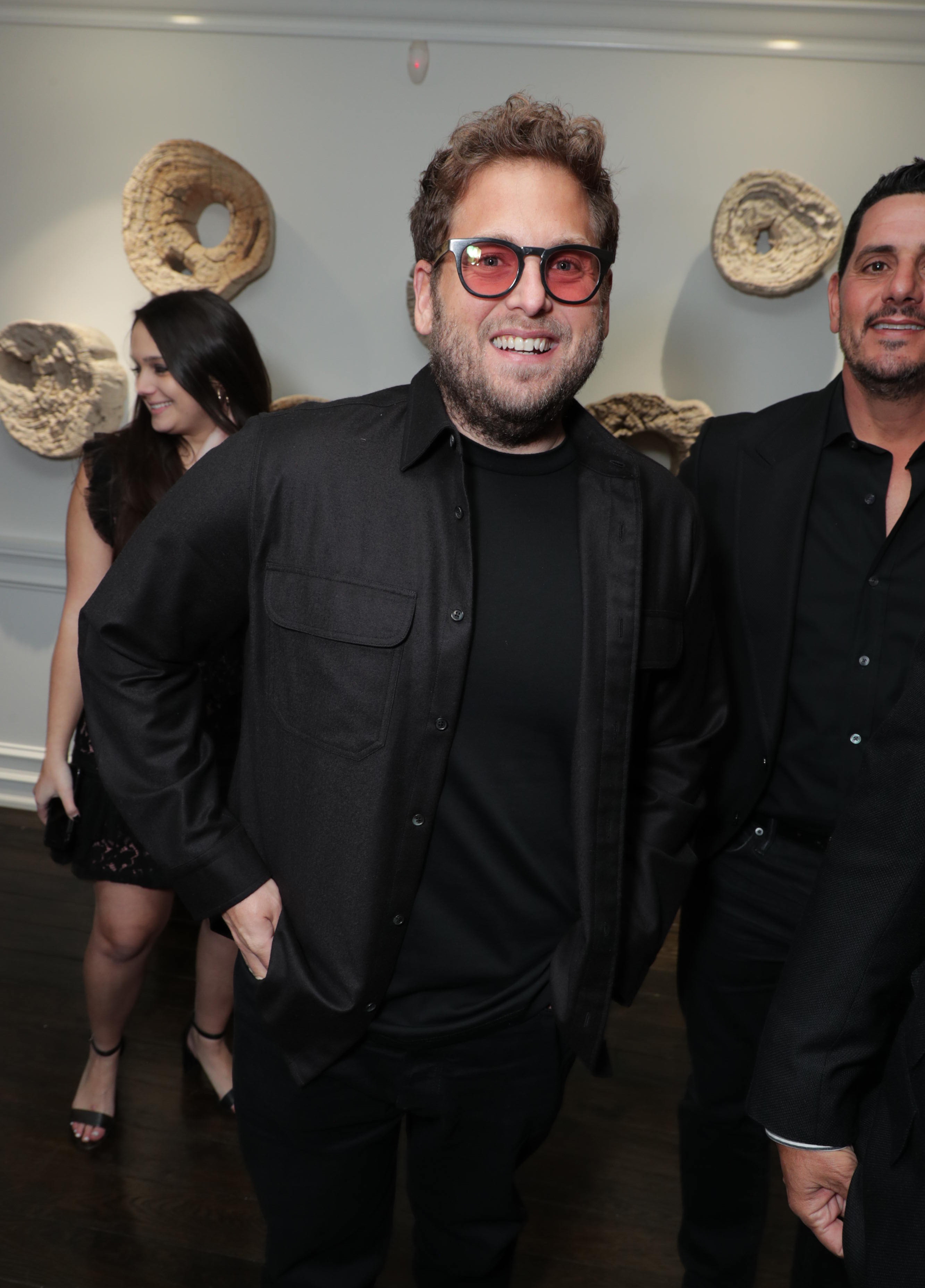 Jonah Hill attends an event in an all-black ensemble, pairing a casual button-up overshirt with a crew-neck tee and tinted glasses. Smiling for the camera, he stands among guests in a warmly lit room with abstract wall art behind him.