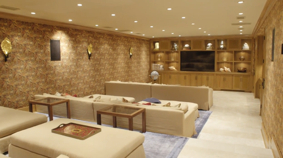 Looking back across the multi-tiered seating layout, plush cream sofas accented with coral and patterned throw pillows line the spacious theater room. Intricate patterned wall coverings, travertine stone flooring, and warm wall sconces complete the luxurious entertainment venue. | Source: YouTube/Architectural Digest
