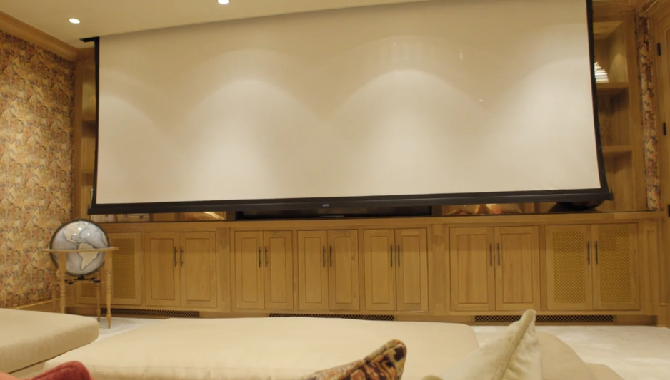 A massive motorized projection screen lowers in front of custom light wood cabinetry, transforming the space into a true cinematic screening room. Soft recessed ceiling lighting illuminates the stage area and plush seating in front of the screen. | Source: YouTube/Architectural Digest