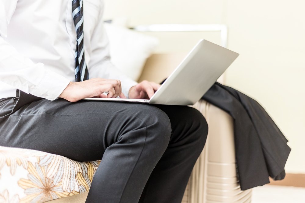 A businessman working on a laptop while sitting on a bed with his travel bag and jacket ready | Photo: Shutterstock/Yuttana Jaowattana