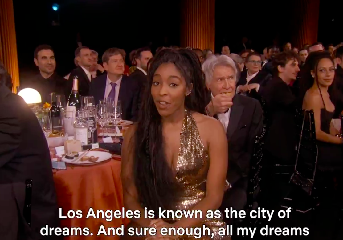 Jessica Williams presenting her speech as Harrison Ford eats and looks at something outside of the camera's view. | Source: X/@netflix