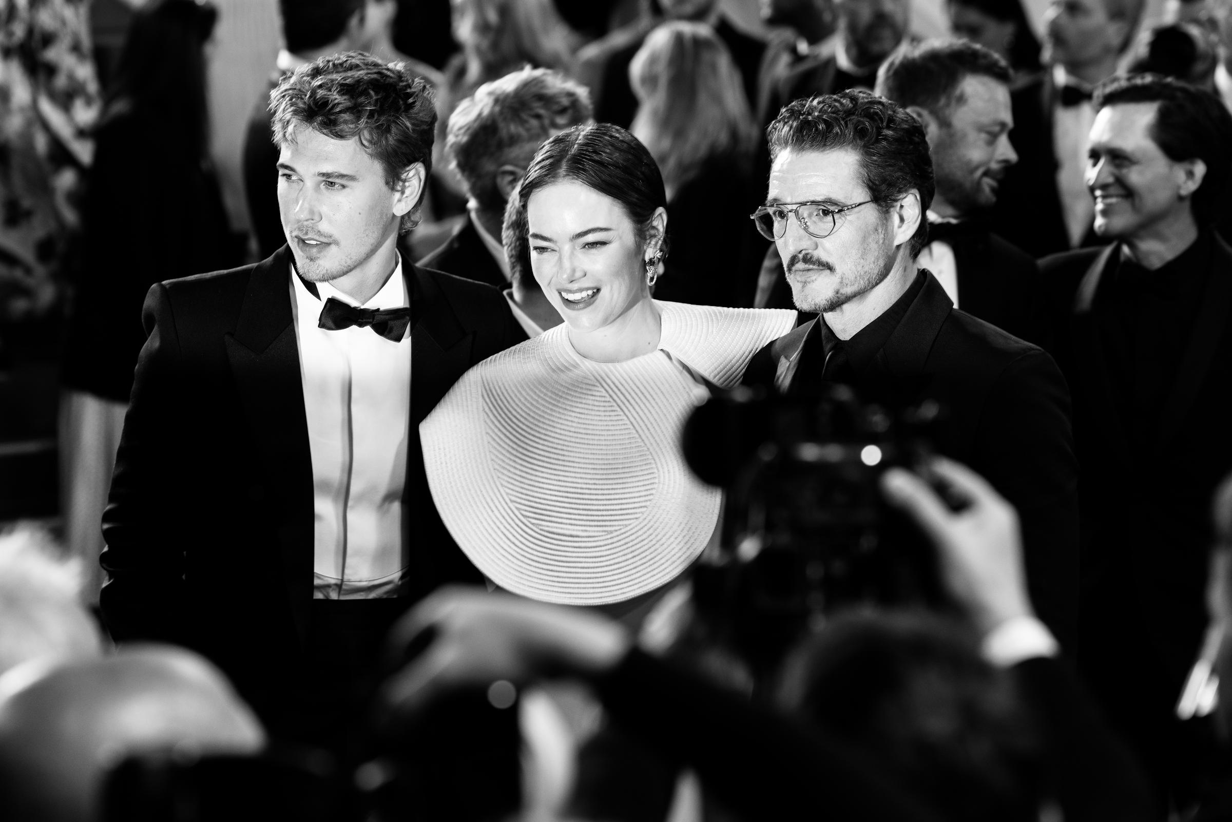 Austin Butler, Emma Stone, and Pedro Pascal pose for photographers at the premiere of "Eddington," captured here in a striking black-and-white moment. The trio exude modern glamour — Stone in a sculptural white gown, flanked by her co-stars in classic tuxedos.