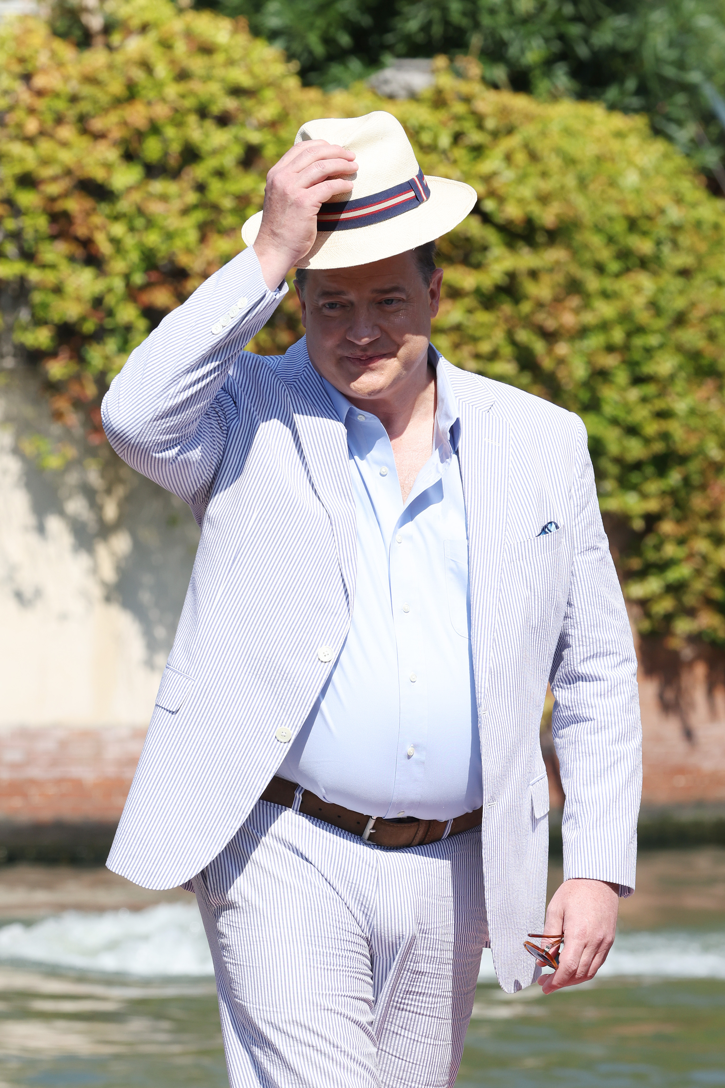 Brendan Fraser tips his cream hat while wearing a pale striped suit and blue shirt during the 79th Venice International Film Festival in Venice, Italy.