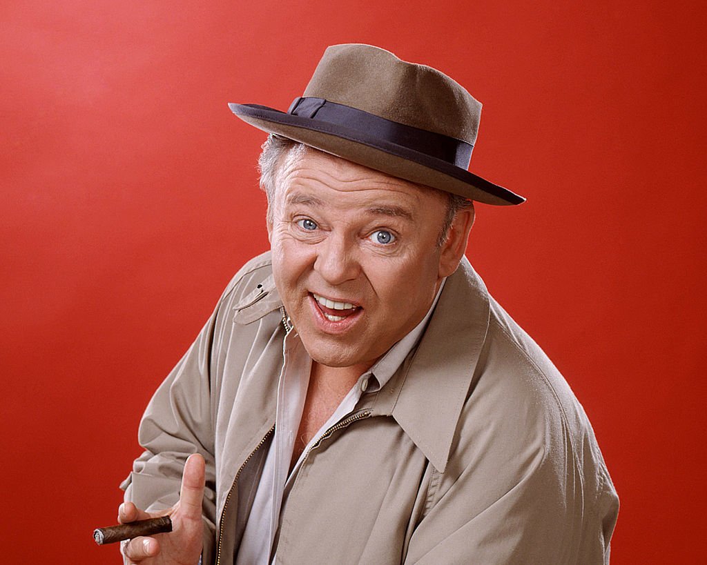 US actor Carroll O'Connor wearing a brown hat with a dark blue band and a beige jacket, and holding a cigar, in a studio portrait circa 1975. | Photo: Getty Images