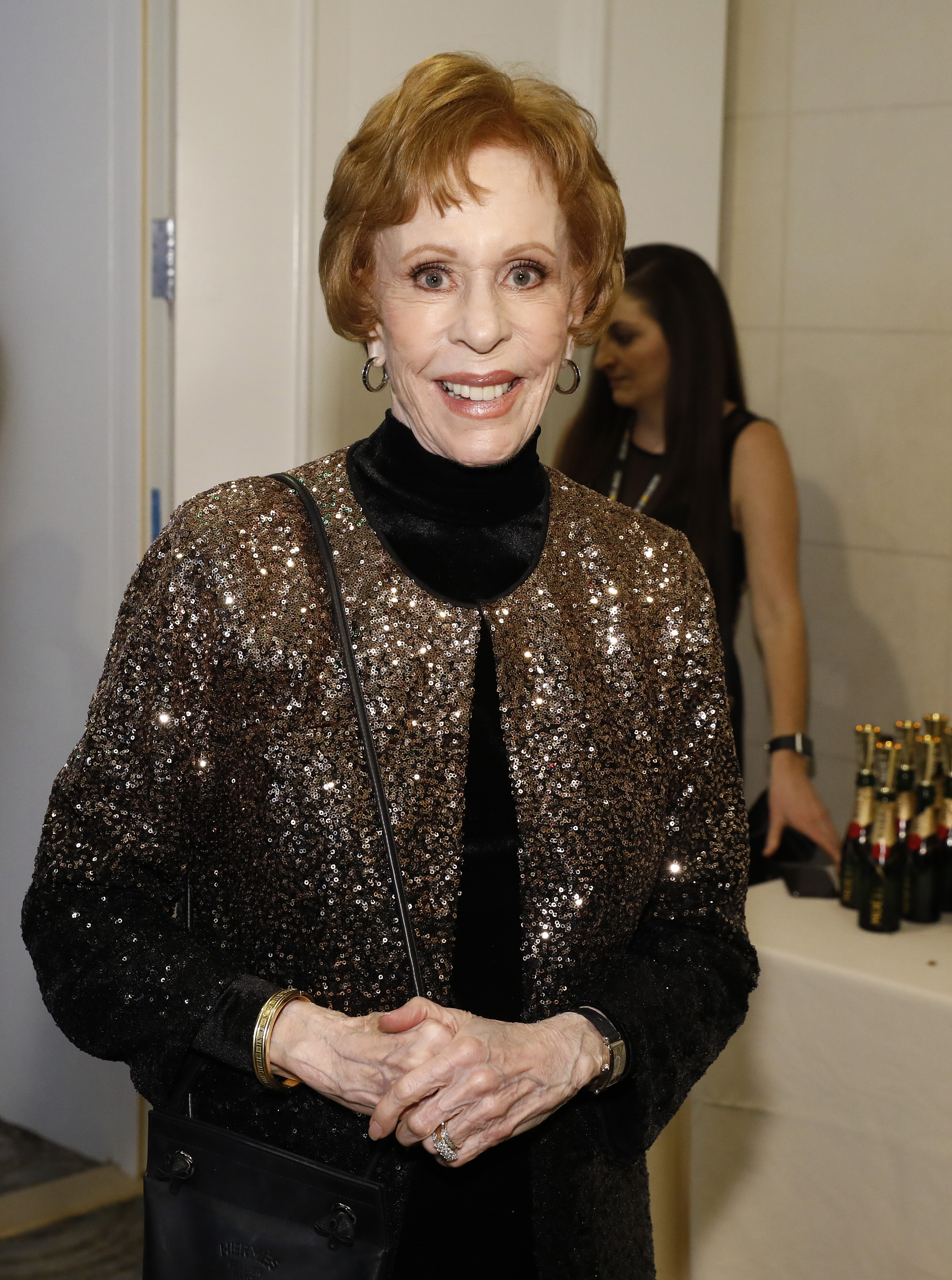 She appears in the press room wearing a black ensemble with a high neckline and a glittering sequined jacket. She stands upright, facing the camera, holding a statuette in both hands. The backdrop behind her features event branding and logos.