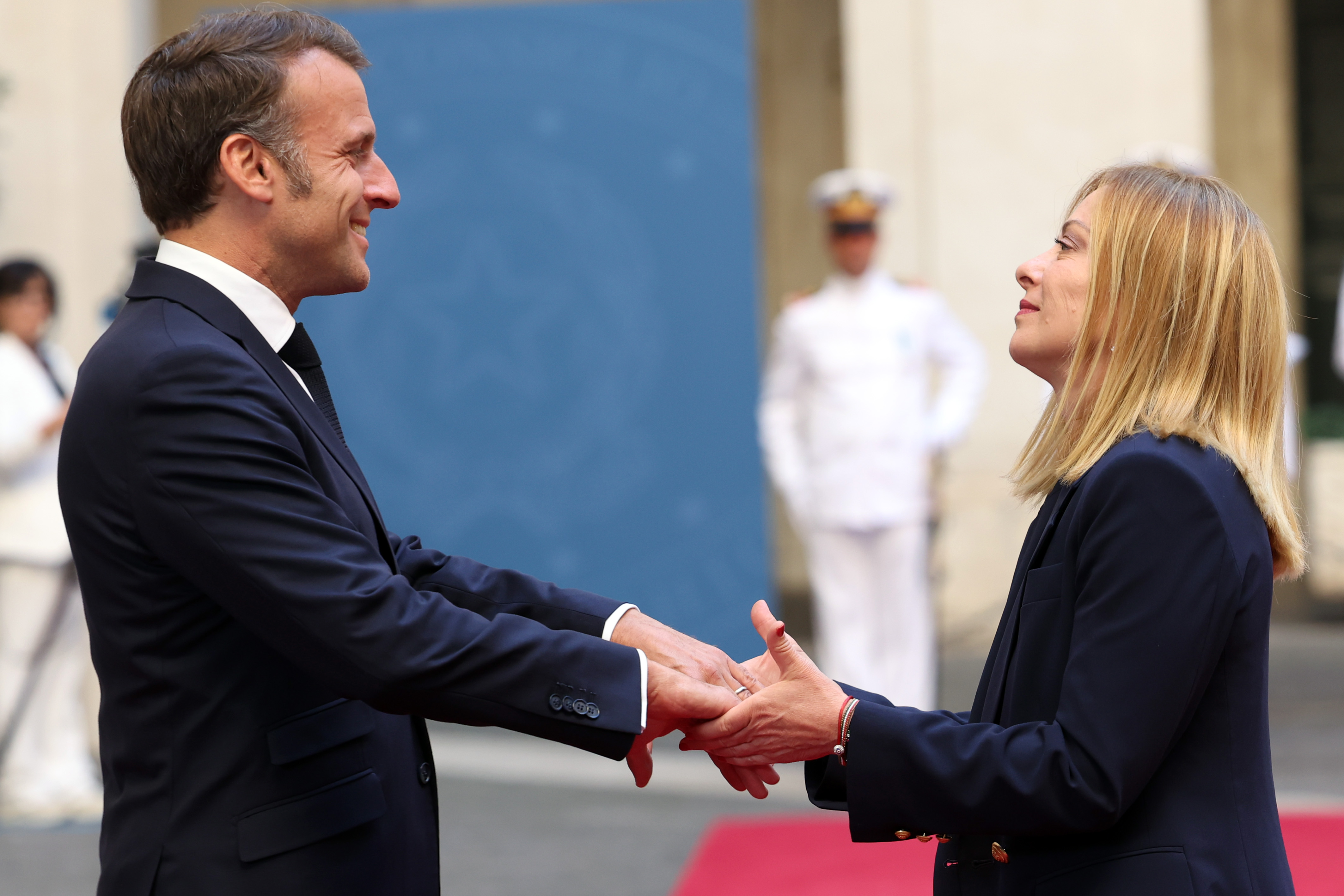 Emmanuel Macron and Giorgia Meloni are caught mid-handshake on a red carpet, with both leaders holding eye contact. Macron smiles broadly while Meloni maintains a composed expression, set against a backdrop of uniformed officials.