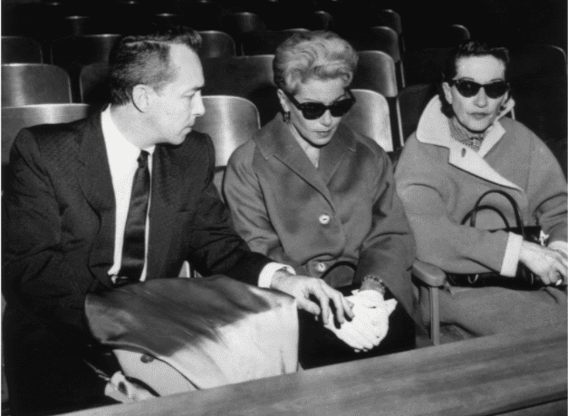 Die amerikanische Schauspielerin Lana Turner (C), die eine dunkle Sonnenbrille trägt, sitzt während des Mordprozesses gegen ihre Tochter Cheryl Crane neben ihrem Ex-Mann Stephen Crane in einem Gerichtssaal. | Quelle: Getty Images