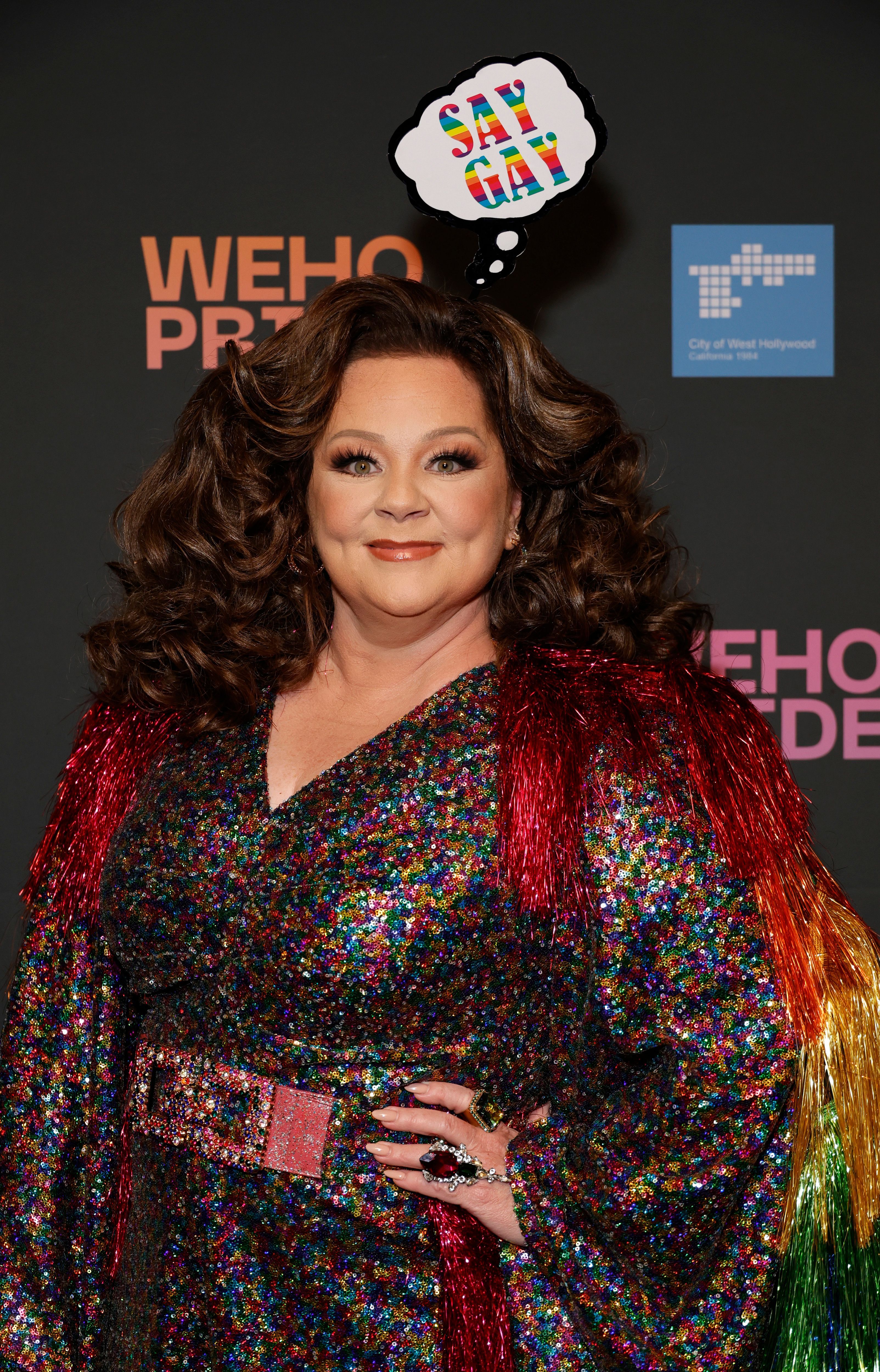 Melisa McCarthy shines in a dazzling rainbow-sequin outfit with bold, voluminous hair and vibrant fringe that celebrates the spirit of Pride. With a playful "Say Gay" thought bubble above her, she radiates joy and unapologetic pride on the WeHo event.