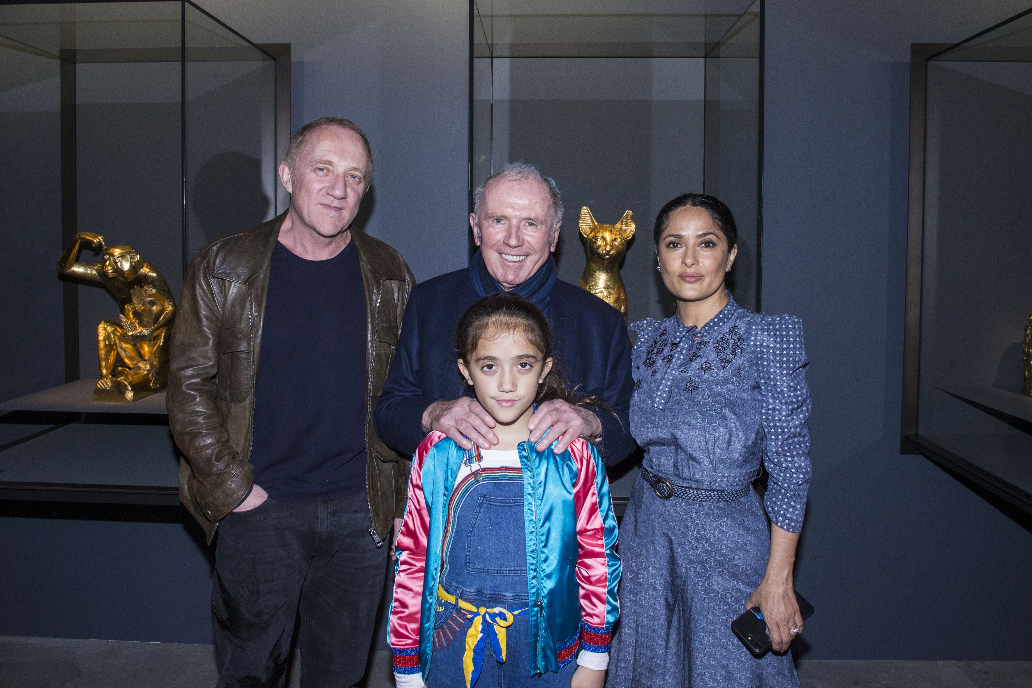 While attending Damien Hirst's exhibition at the Venice Biennale, Valentina Pinault poses with her mother Salma Hayek, father François-Henri Pinault, and grandfather François Pinault. Set against a modern gallery backdrop, Valentina sports a bright satin bomber jacket and overalls, standing confidently in front of the adults.
