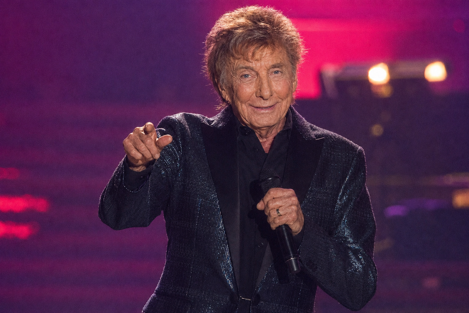 This final image brings the comparison back to the stage, where Manilow has spent much of his career. His familiar microphone, sparkling jacket, and performance pose remain unchanged, making the imagined older face the main difference viewers notice. | Source: ChatGPT