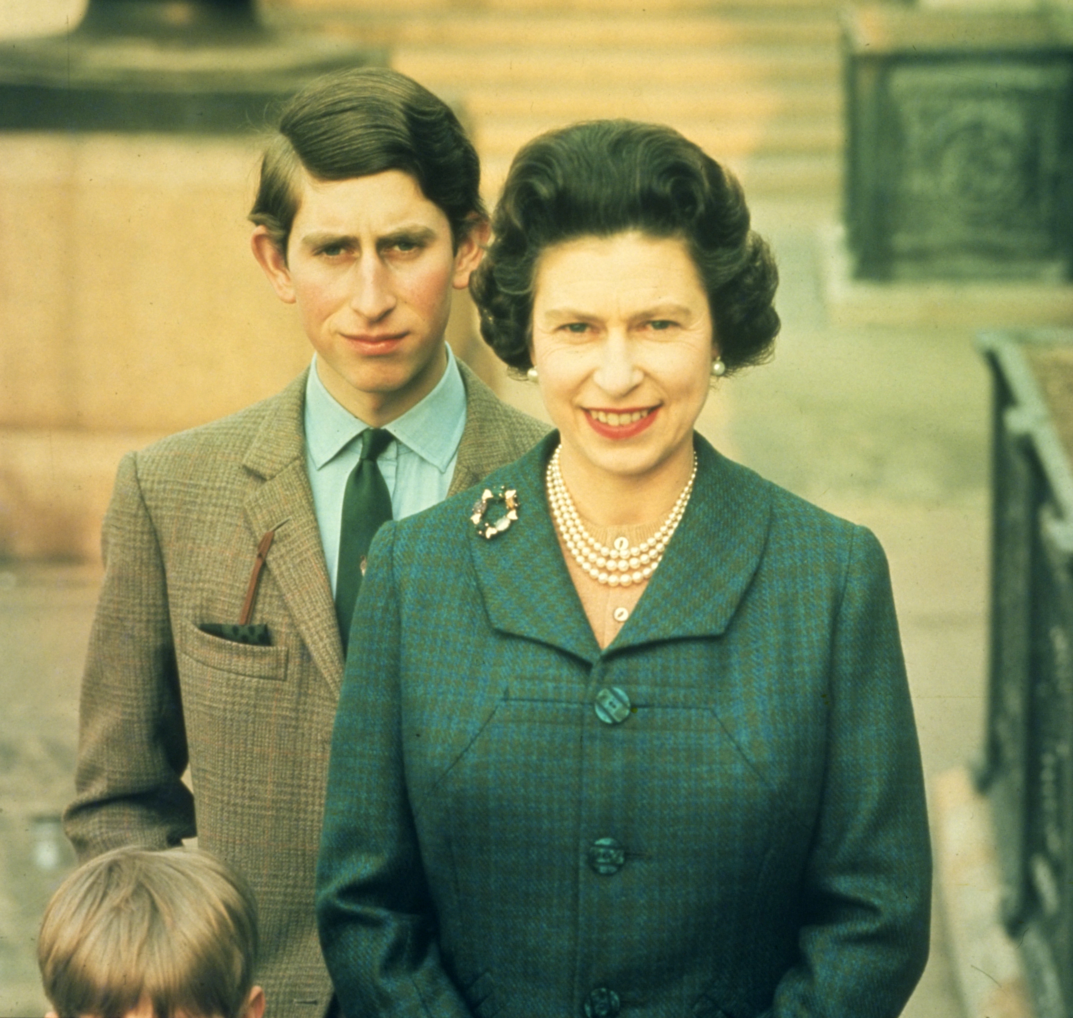 In a poised portrait taken at Windsor Castle in April 1969, Queen Elizabeth II stands elegantly in the foreground, her signature pearls and composed smile reflecting quiet confidence, while Charles, Prince of Wales, appears just behind her, offering a more reserved presence as the image captures a moment of transition between generations within the royal family.