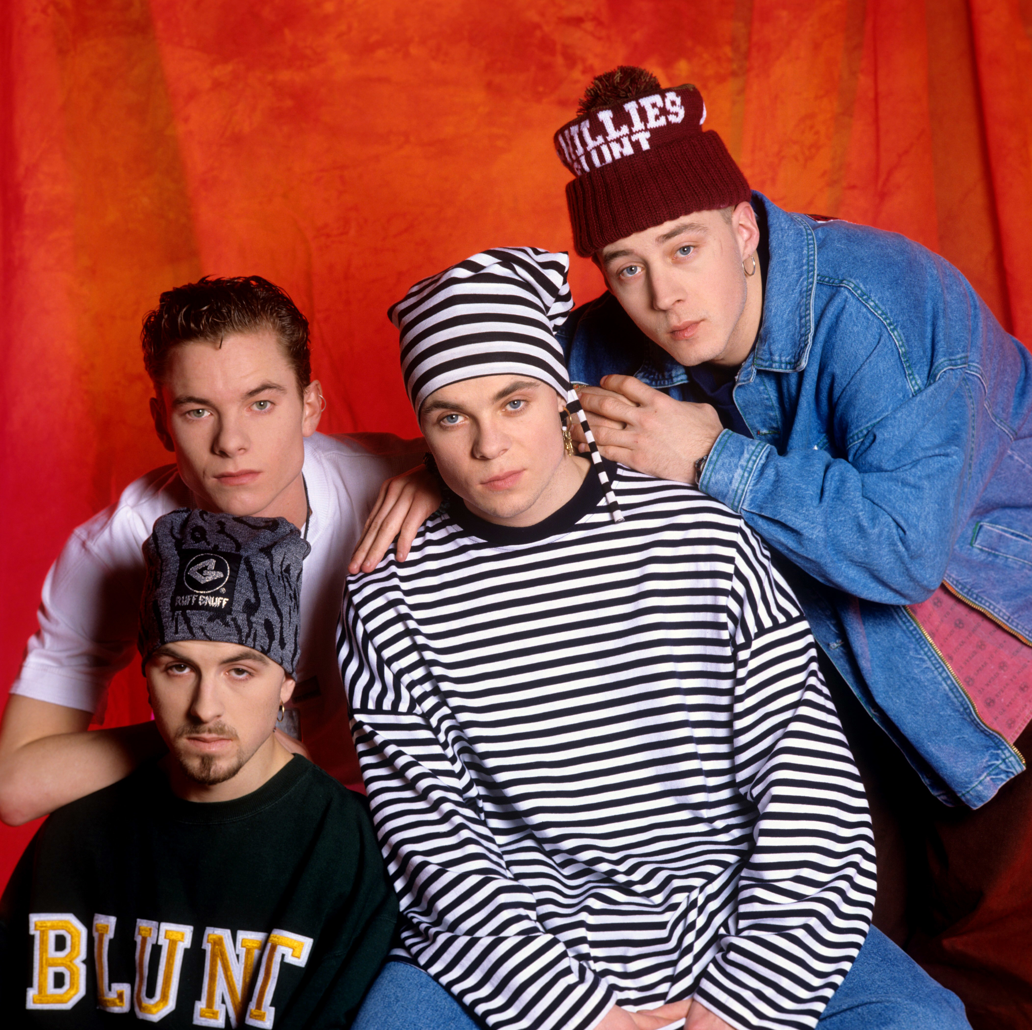 In this striking studio portrait, Terry Coldwell, Tony Mortimer, Brian Harvey, and John Hendy lean into their distinctive aesthetic, pairing bold stripes, oversized denim, and statement headwear with unwavering, almost confrontational expressions. The vivid backdrop heightens the intensity of the shot, capturing the group's edgy image and the quiet confidence that set them apart during their ascent in the 1990s pop landscape.