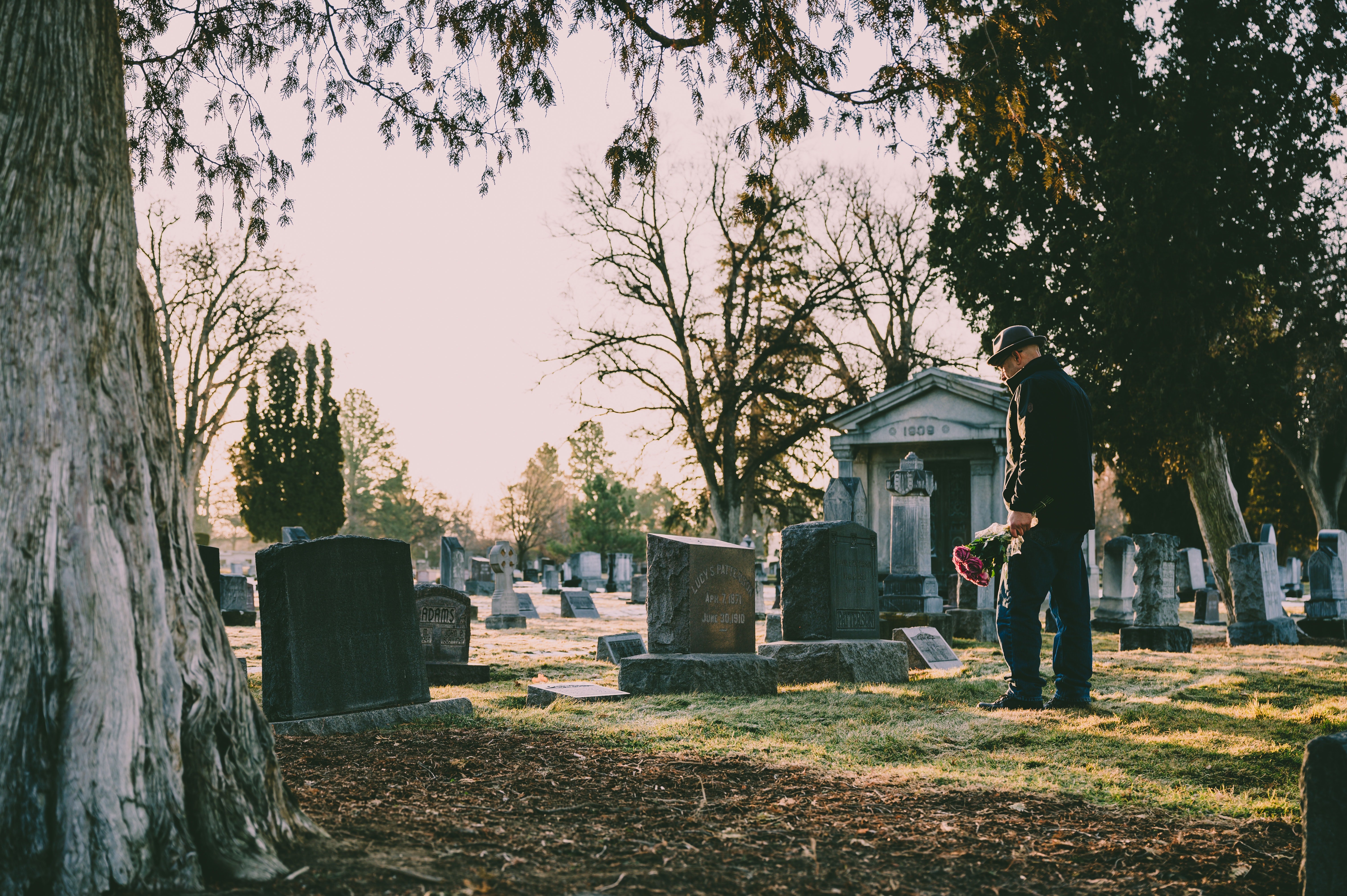 Man standing in a black jacket in front of a grave with flowers in his hand. | Photo: Pexels