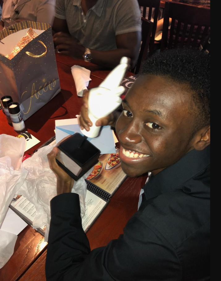 Beaming with excitement at a restaurant table, Donovan Metayer holds up a small gift box while clutching what appears to be a celebratory item, his wide smile capturing a joyful, intimate moment. | Source: Gofundme