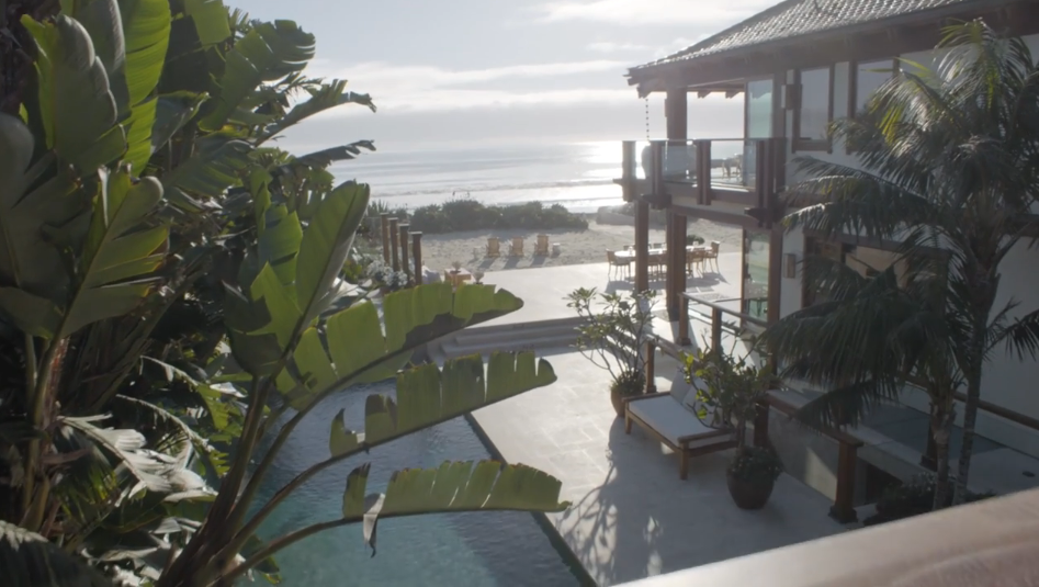 A view from behind vibrant banana leaves frames the saltwater pool and stone patio stretching out toward the beach. The multi-level rear exterior of the home, complete with wooden balconies and dining decks, overlooks the ocean horizon under a soft coastal sky. | Source: YouTube/Architectural Digest