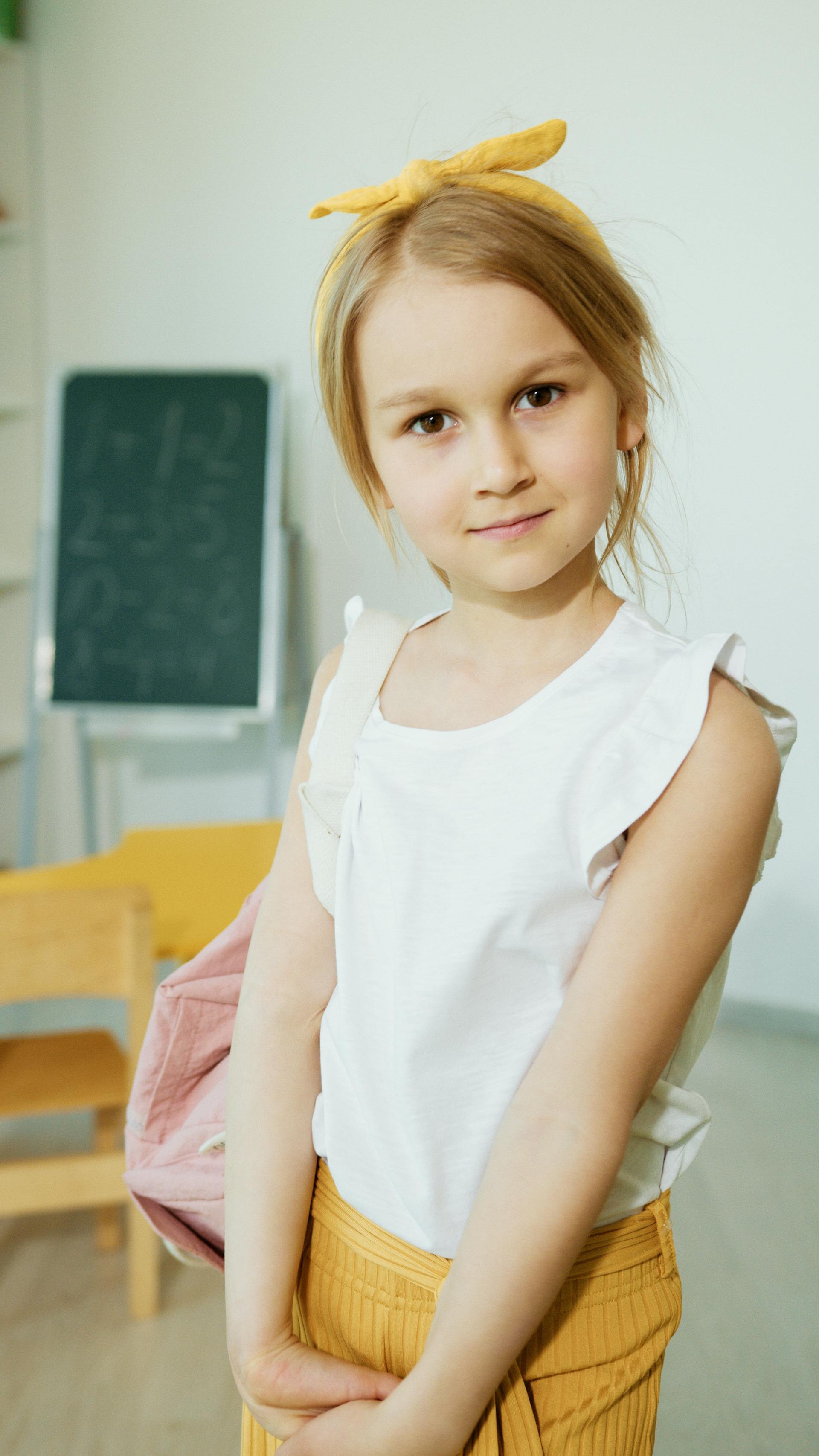 A little girl in a white sleeveless blouse smiling in a room | Source: Pexels