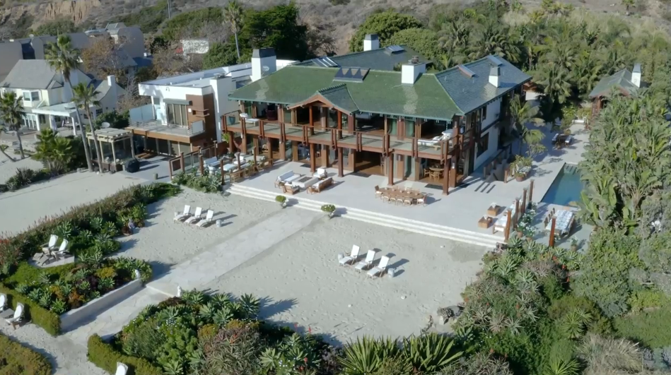 A bird’s-eye view captures the grand rear exterior of Orchid House, showcasing its green tiled roof, expansive wooden balconies, and multi-level outdoor seating areas. The property opens directly onto a private sandy courtyard surrounded by lush tropical landscaping, with a swimming pool tucked away to the side. | Source: YouTube/Architectural Digest